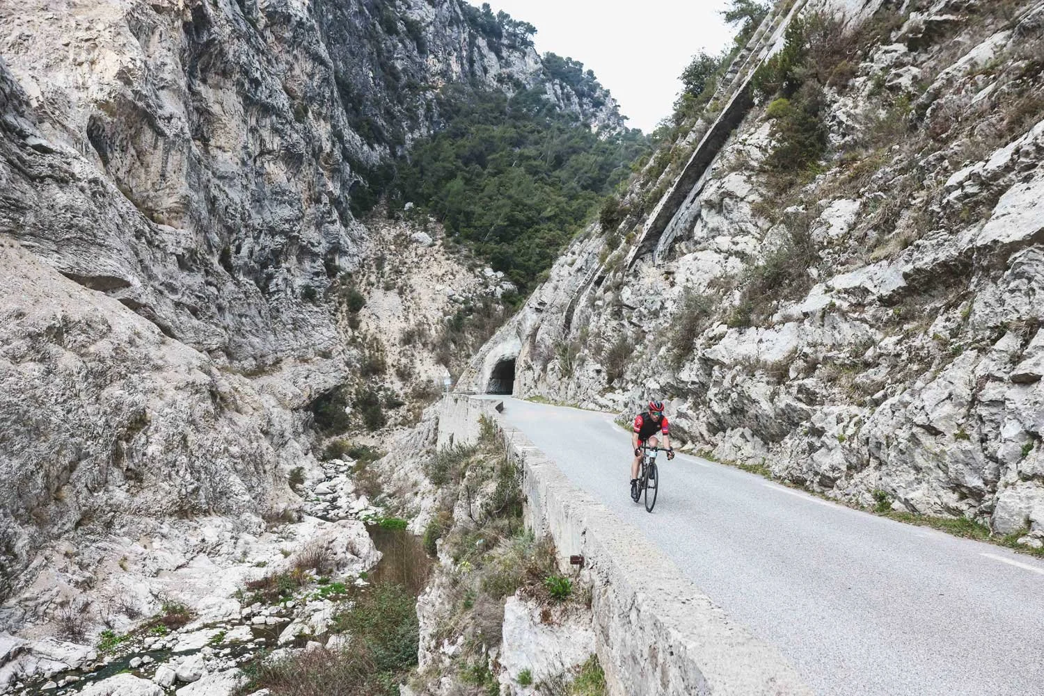 A cyclist wearing a red and black outfit riding a mountain bike along a narrow mountain road with rocky cliffs and trees surrounding the path.