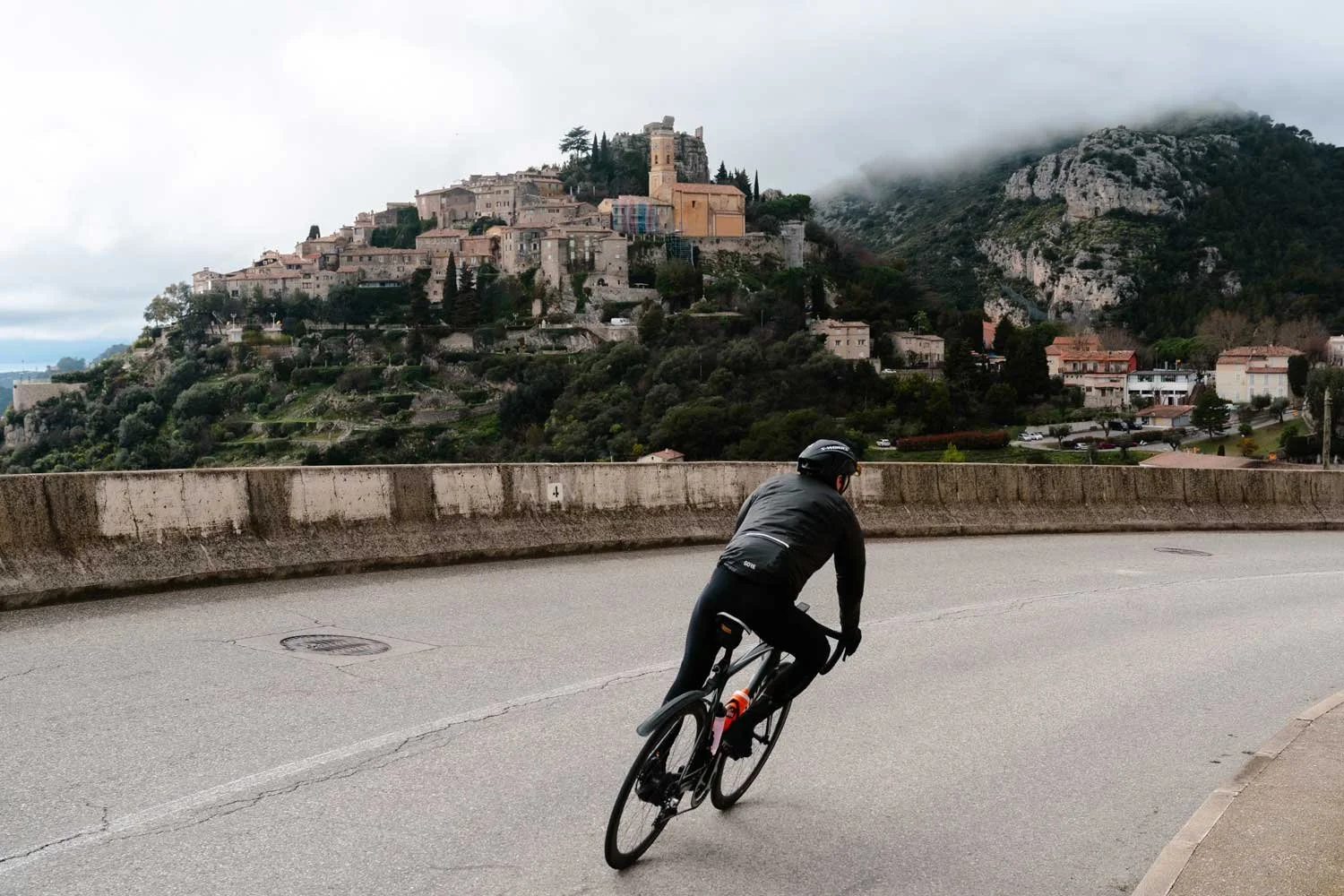A person riding a bicycle on a winding road with a hilltop medieval town and fortress in the background, partly shrouded in fog.