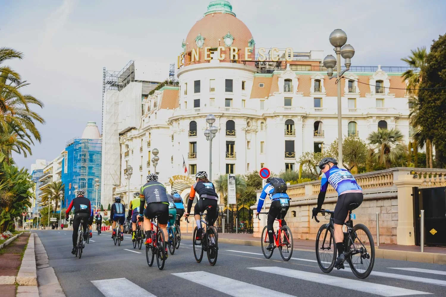 Group of cyclists riding on a city street with a large historic hotel building in the background, palm trees lining the street, and a crosswalk visible.