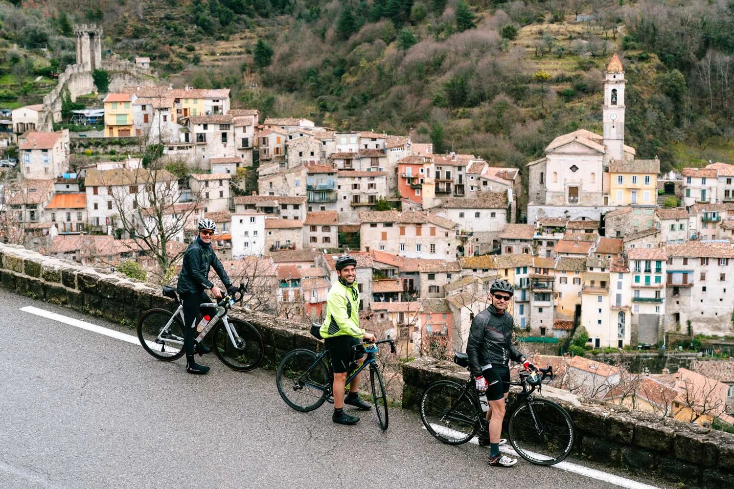 Three men with bicycles on a hillside road overlooking a small town with colorful buildings and a church tower.