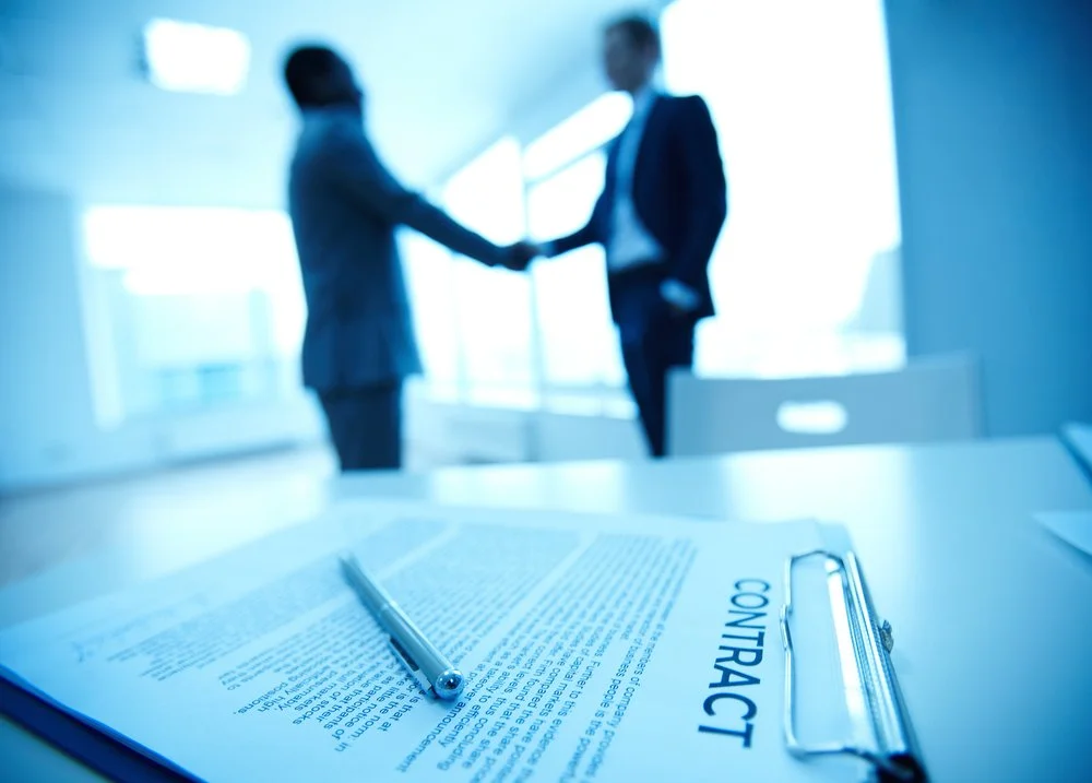 Close-up of a contract and a pen on a table with two business professionals shaking hands in the background, silhouette and blurred.