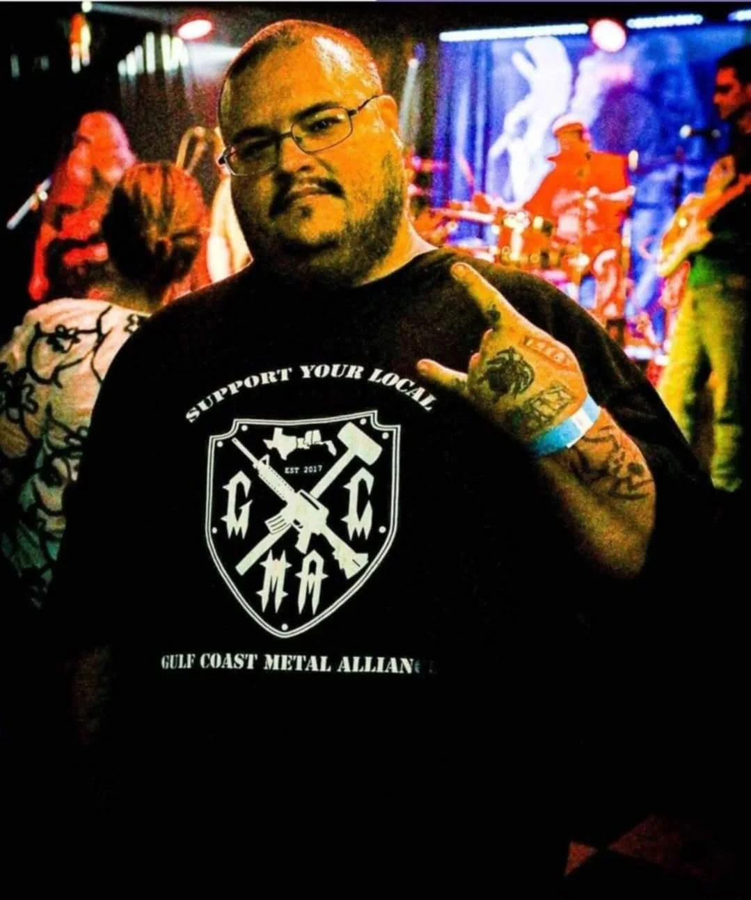 A man with glasses and tattoos wearing a black T-shirt with a metal emblem and the phrase support your local, standing in front of a colorful stage with musicians playing. The stage background includes a large screen displaying a person with raised arms.