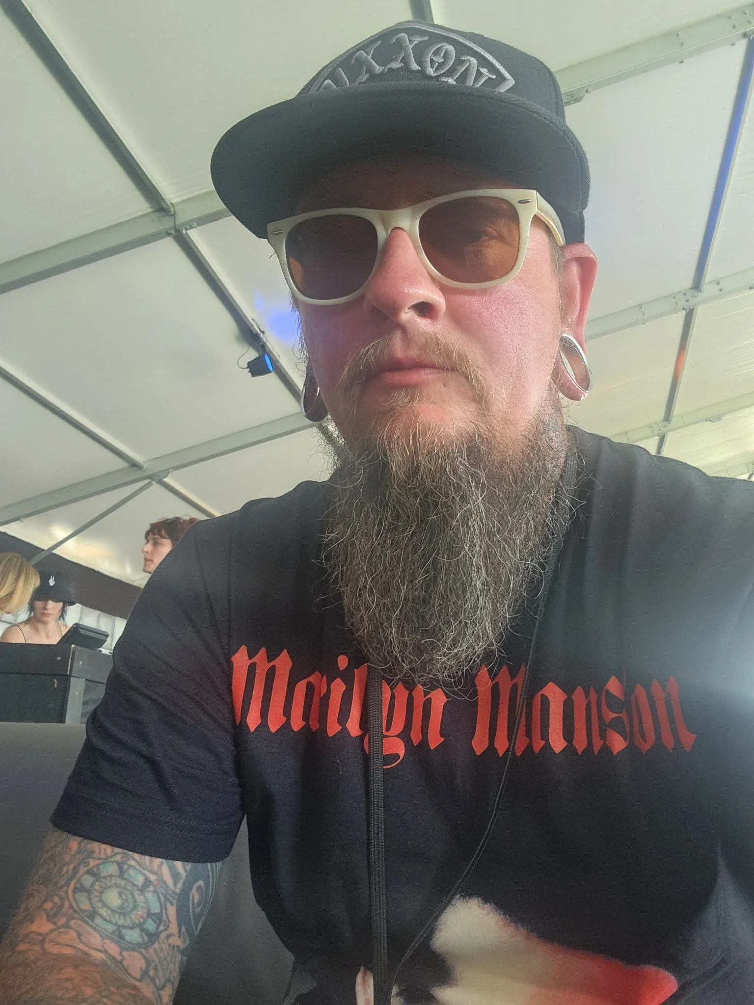 A man with a long beard and tattoos on his arm wearing sunglasses, a black baseball cap with a logo, and a black Marilyn Manson T-shirt taking a selfie inside a tent.