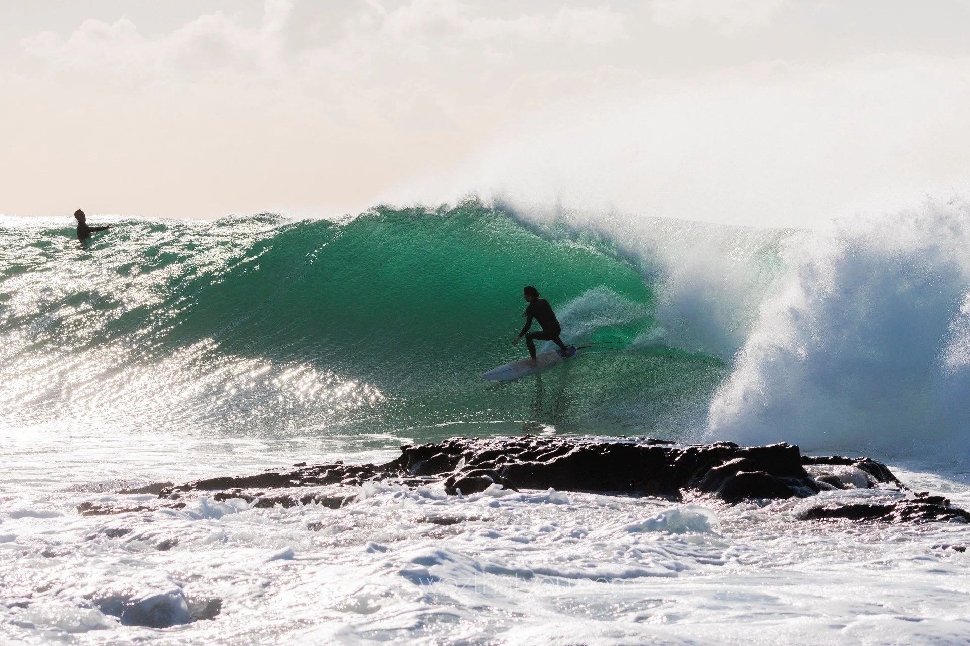 Surfer riding inside a large green wave with a rocky beach in the foreground and another person in the distance