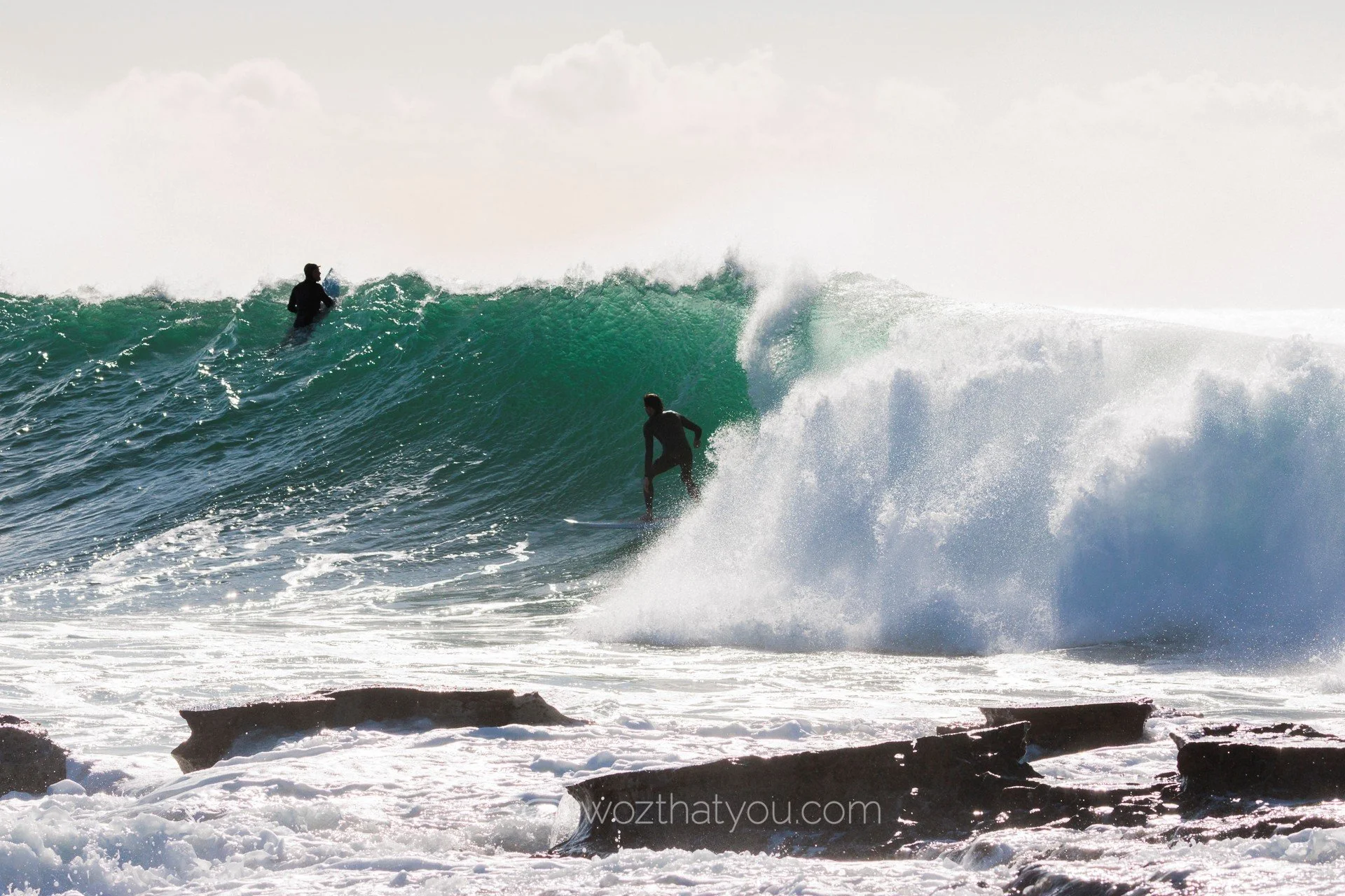 Two surfers riding a large ocean wave with rocks in the foreground.