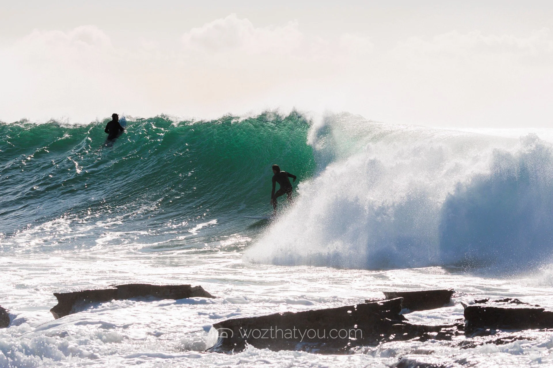 Two surfers riding a large wave near rocky shoreline in the ocean.