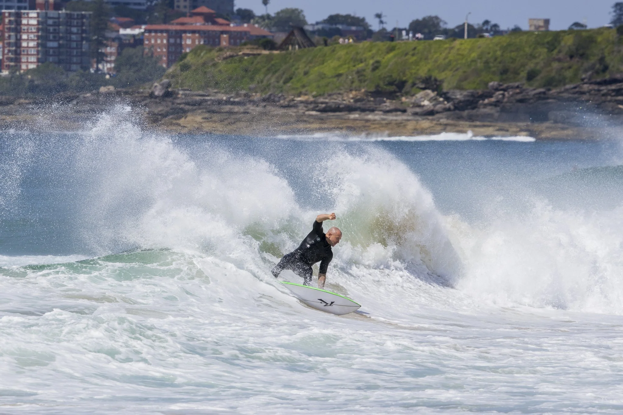 A man surfing on a wave near a coastline with buildings and greenery in the background.