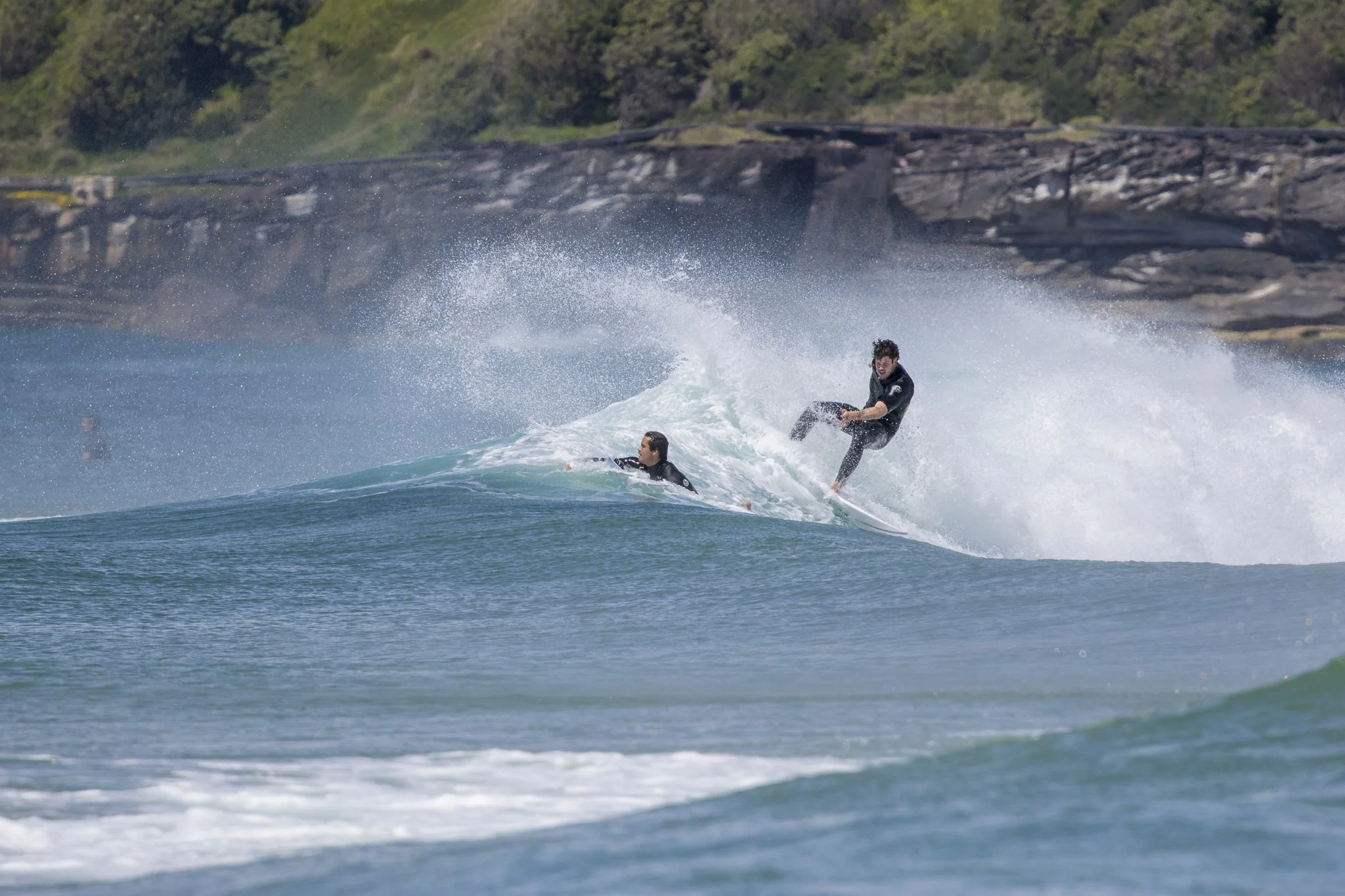 Two surfers riding a wave at the beach with a rocky shoreline and green hills in the background.