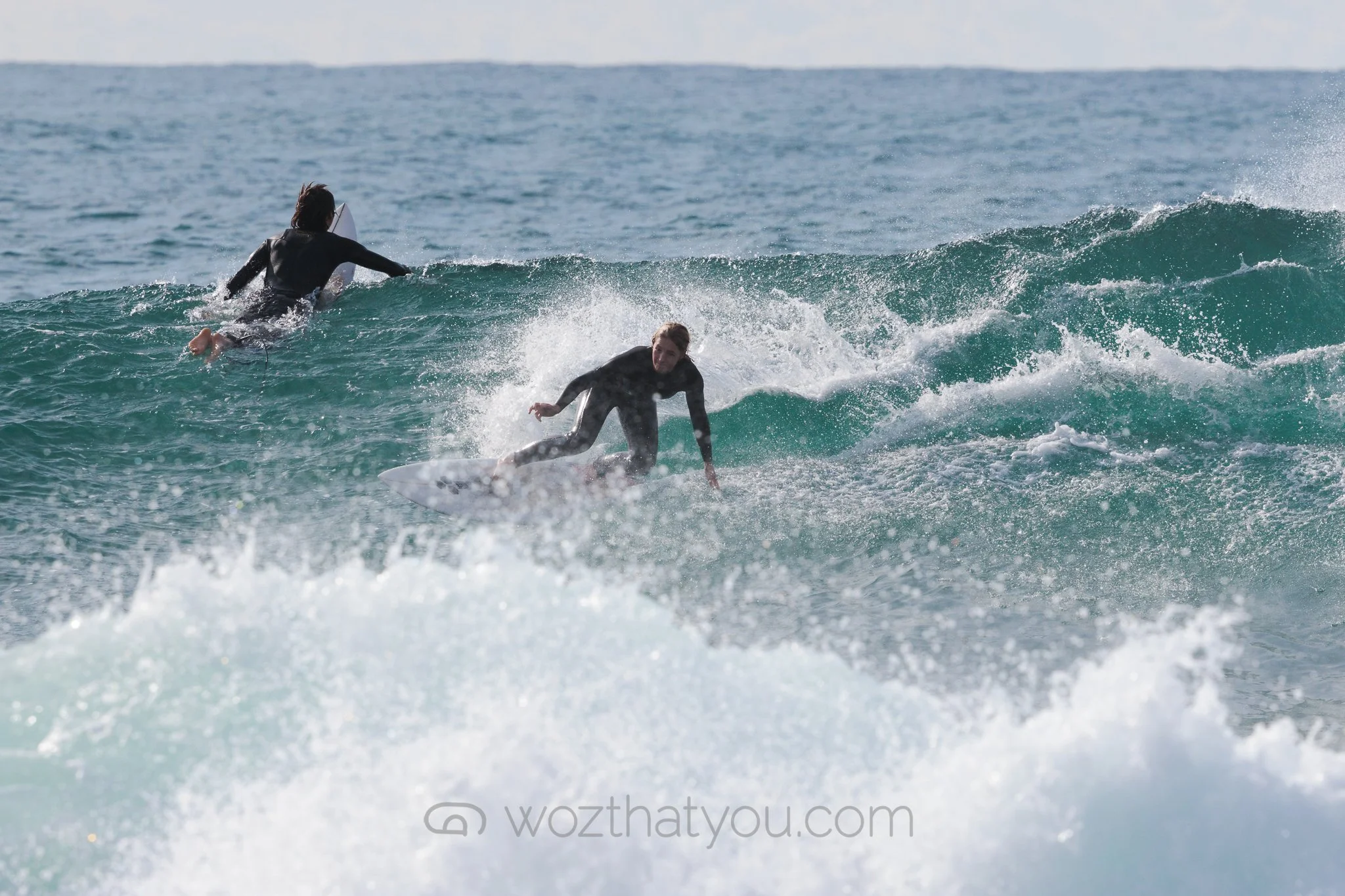 Two surfers in black wetsuits riding on ocean waves, with one falling and the other paddling on her surfboard.