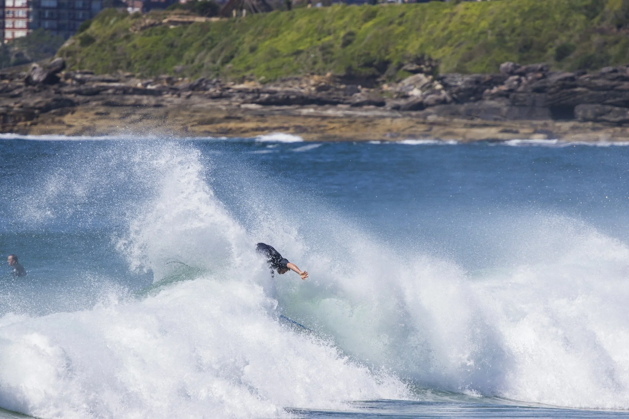 A person surfing on a wave near a rocky coastline with green hills in the background.