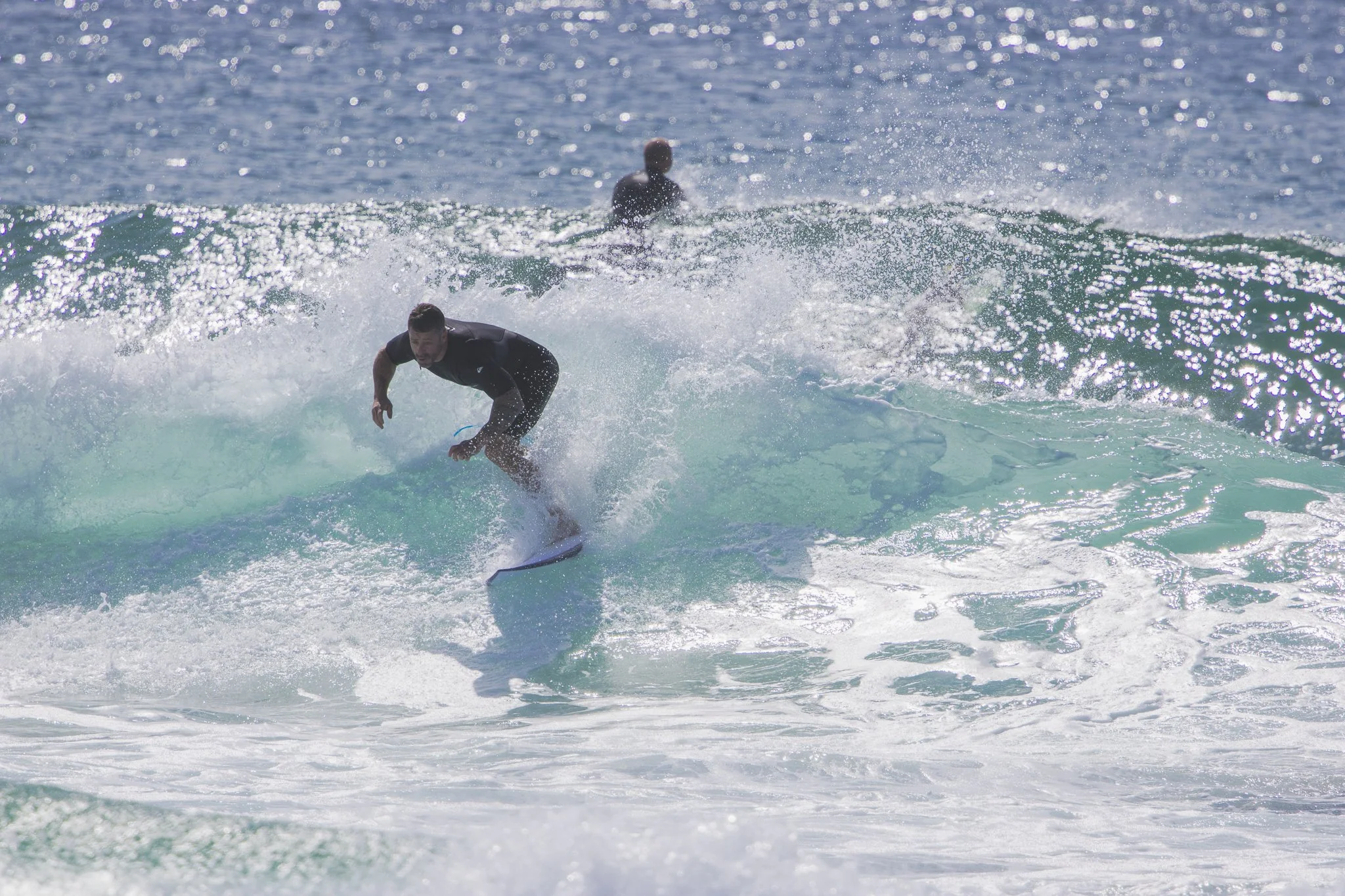 A man in a black wetsuit surfboarding on a breaking wave in the ocean with another surfer in the background.