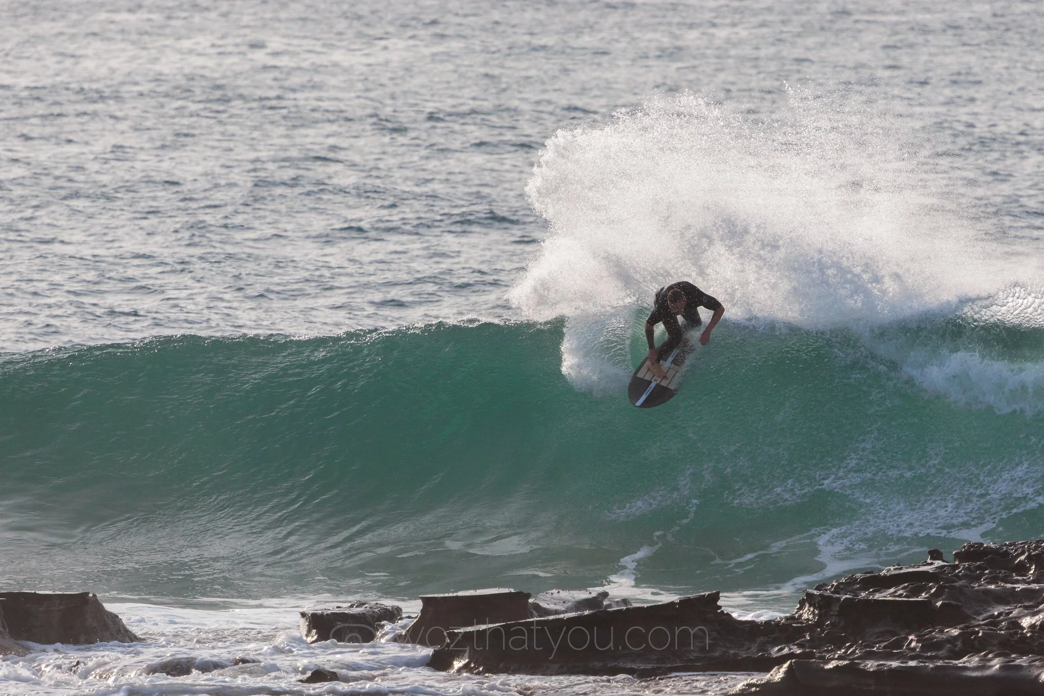 A person surfing on a wave in the ocean, with rocky shoreline in the foreground.