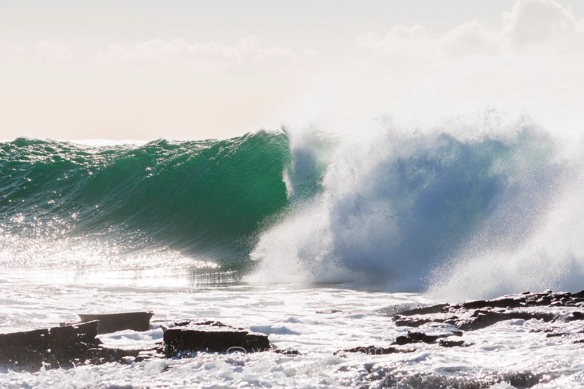 A large green ocean wave crashing onto rocky shore with white sea foam.