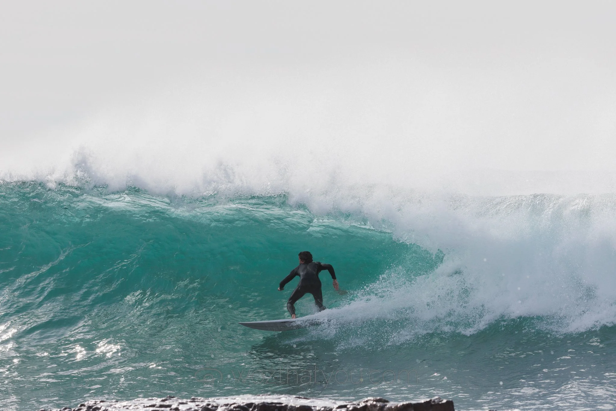 Person surfing on a large ocean wave with rocky shoreline in the foreground.