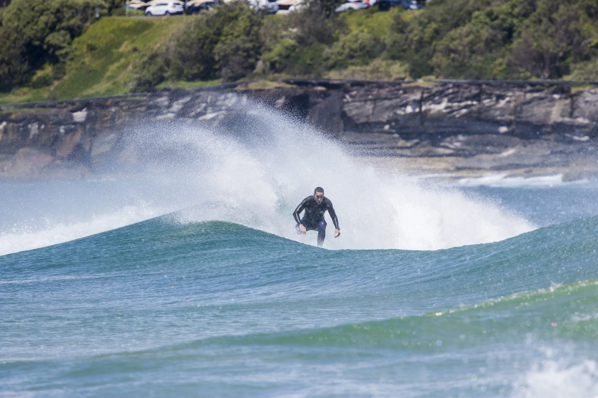 A person wearing a black wetsuit surfing on a wave near the shore with a rock formation and some cars on a hill in the background.