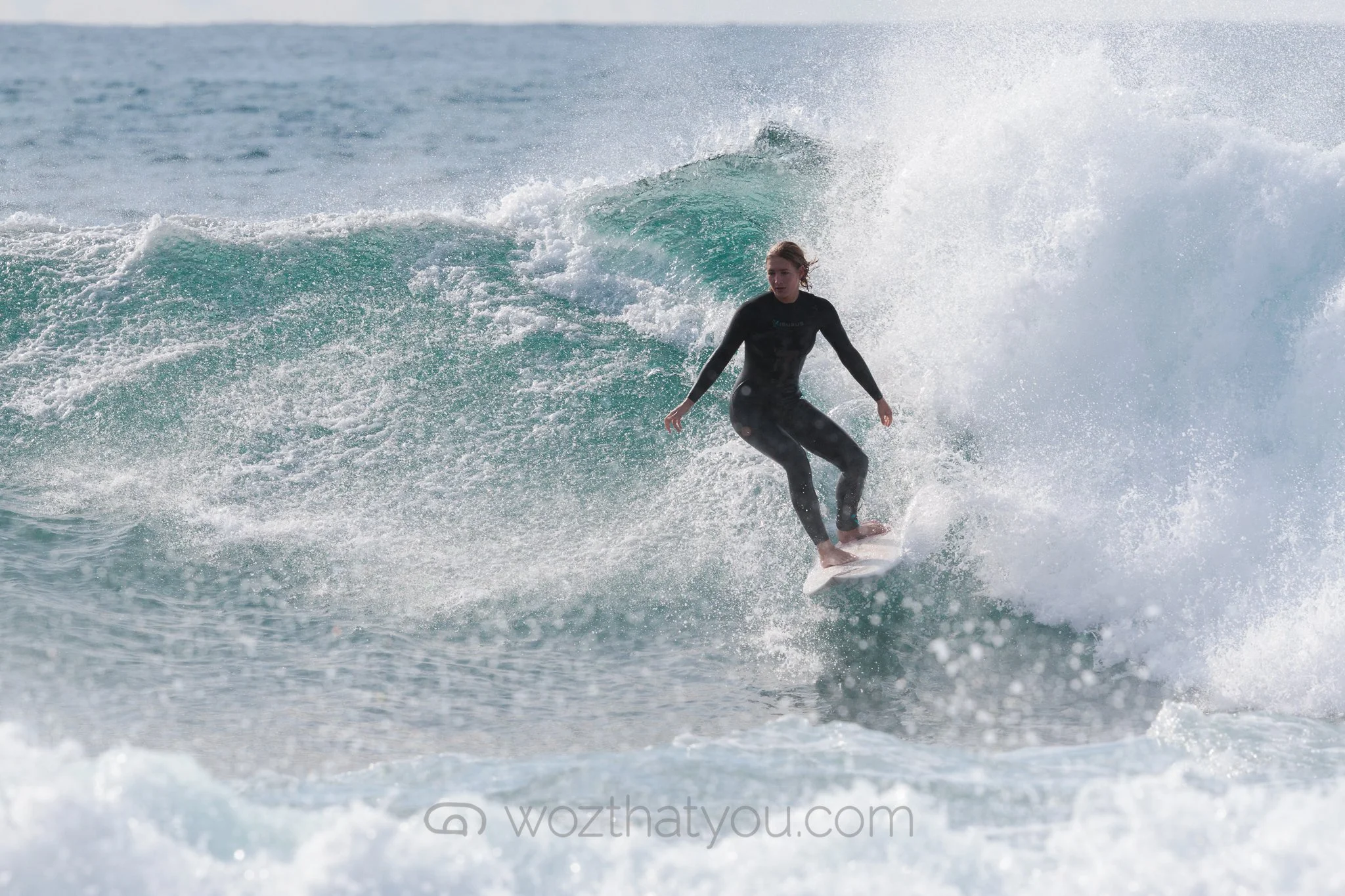 A woman surfing on a wave in the ocean, wearing a black wetsuit, with water splashing around her.
