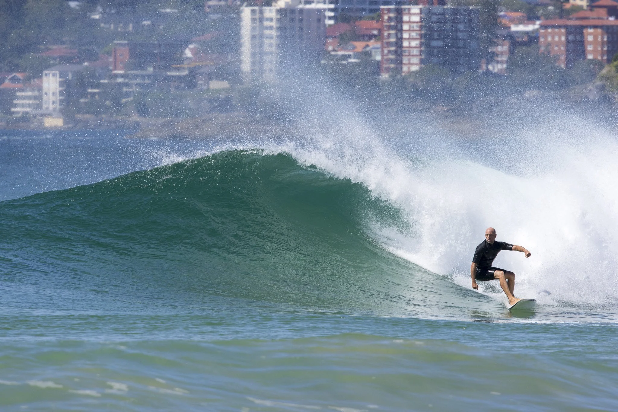 A man surfing on a wave near a city with tall buildings in the background.