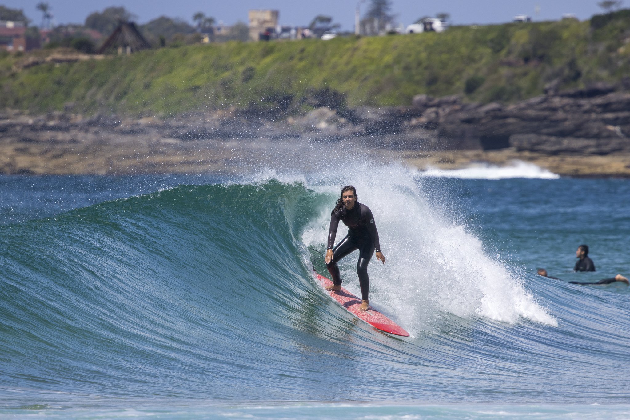 A person in a black wetsuit surfing on a pink surfboard on a blue wave near the shoreline with other surfers in the background.
