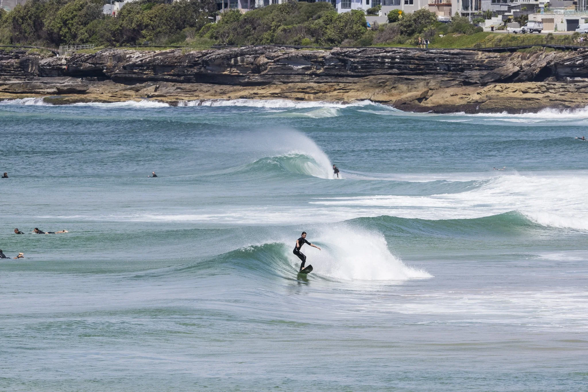 Surfer riding a wave at the beach with several other surfers in the water and a rocky coastline with houses in the background.