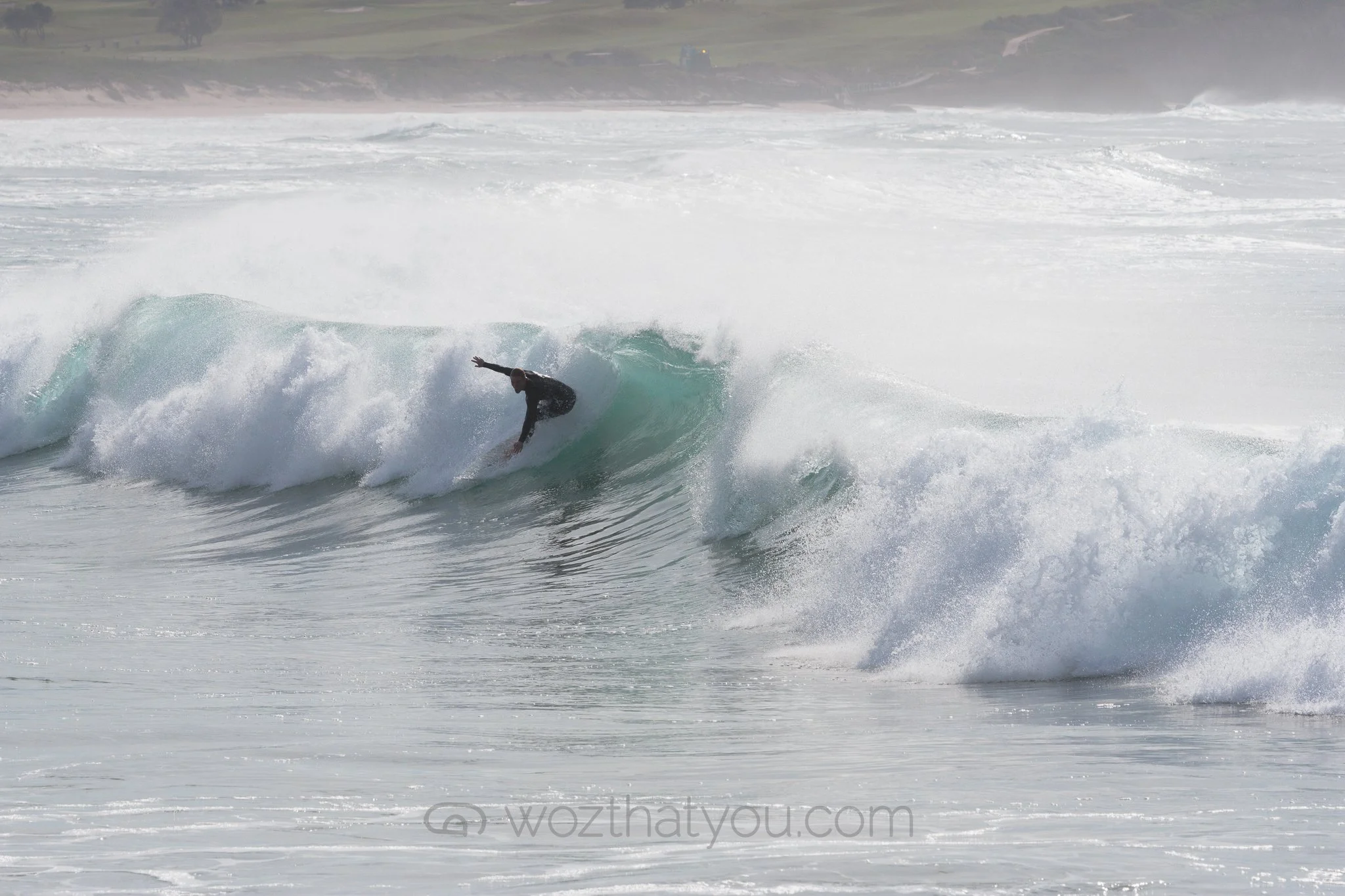 A person surfing on a wave at the beach with a shoreline and distant land in the background.