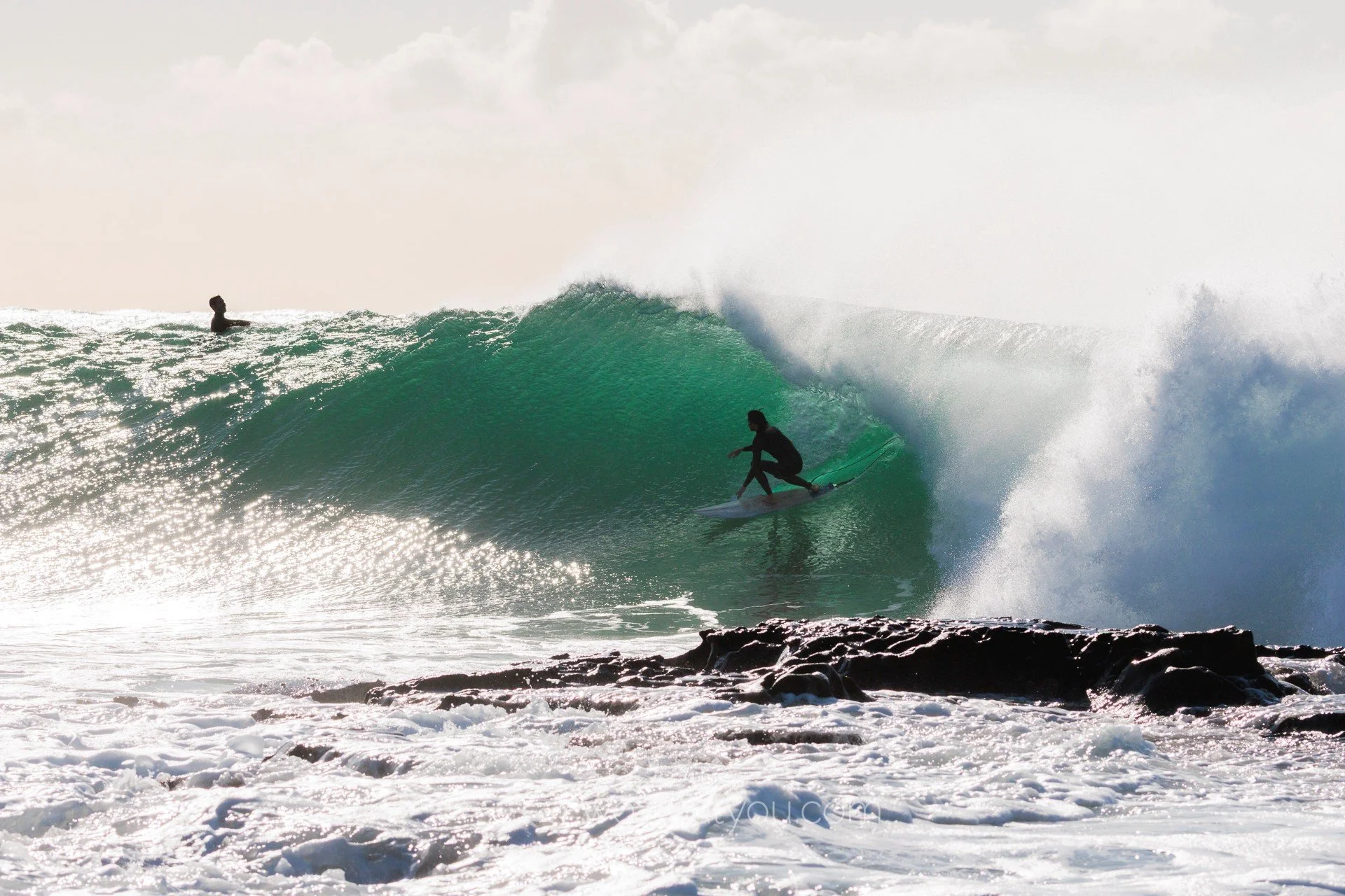 Surfer riding inside a large green wave near rocky shoreline, with another person on the water in the distance.