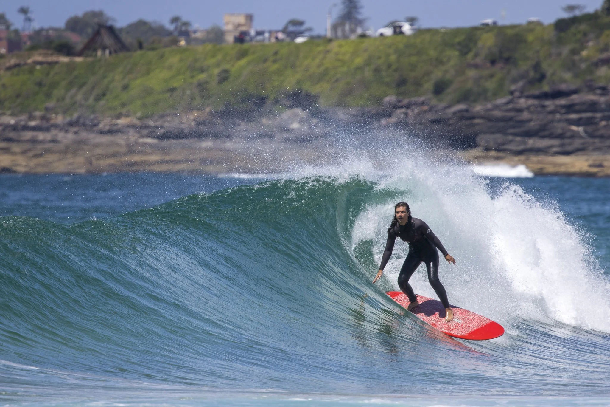 A woman surfing on a red surfboard in the ocean, riding a wave with rocks and a grassy hill in the background.