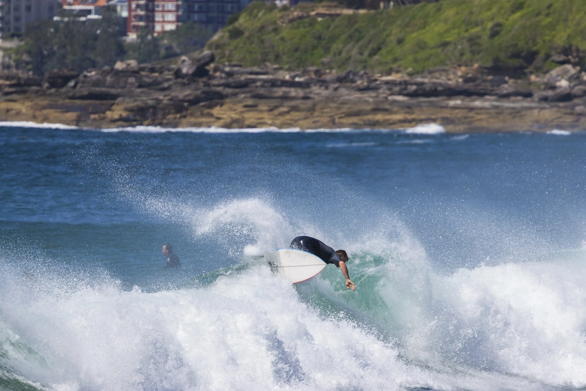 A person surfing on a wave in the ocean with a rocky and green shoreline in the background.
