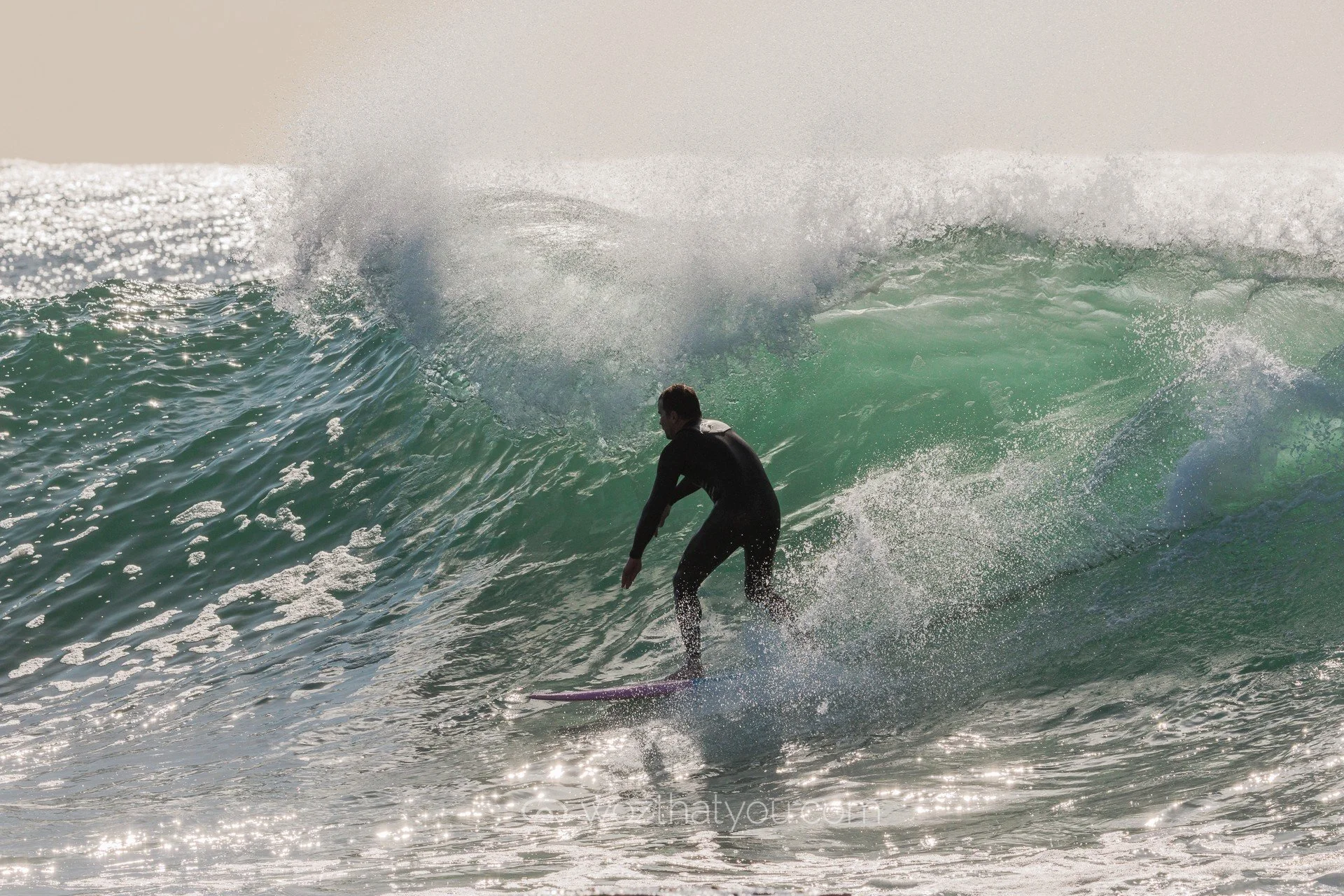 A person in a wetsuit surfing a large ocean wave on a pink surfboard with sunlight reflecting off the water.