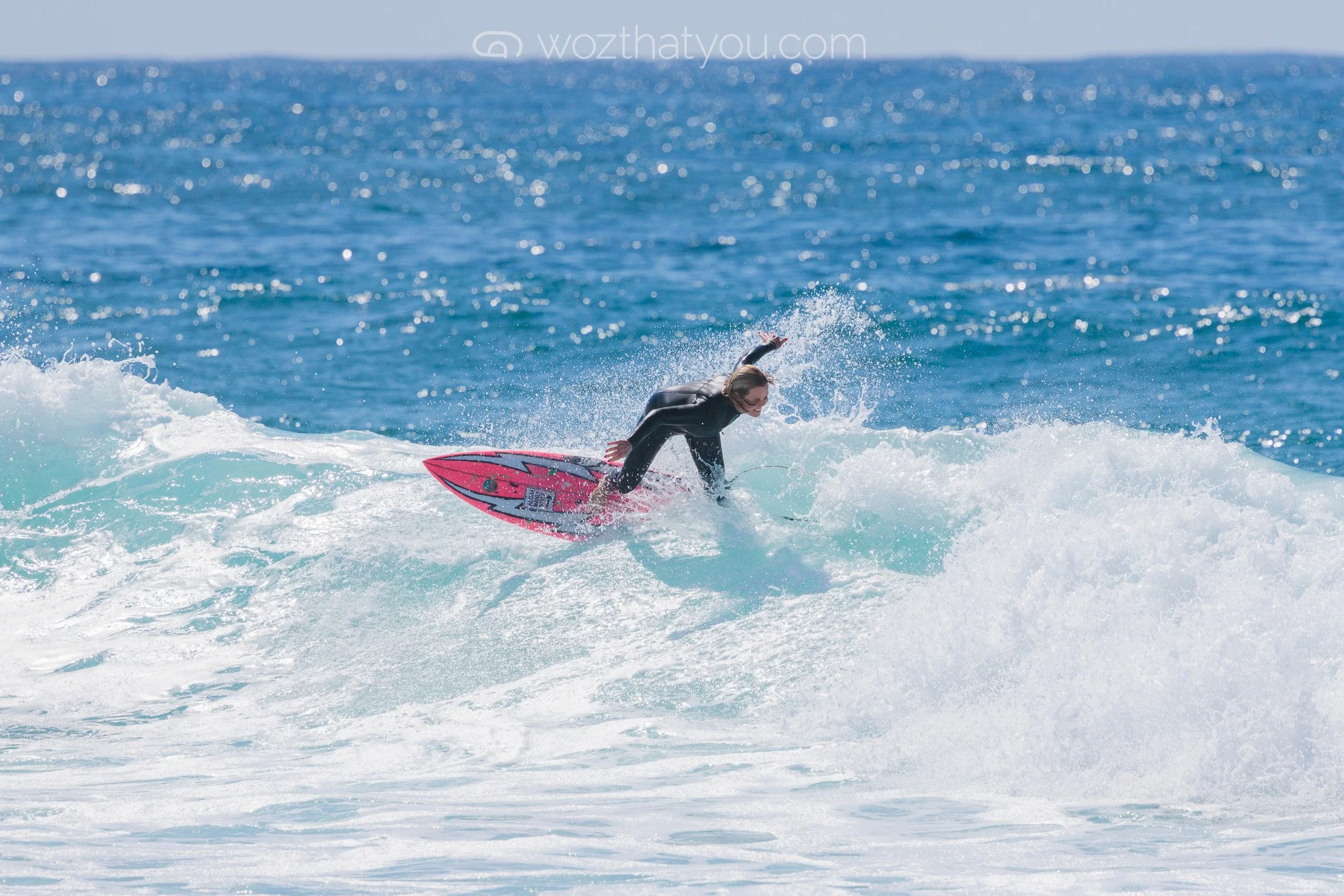 A woman surfing on a pink and black surfboard in the ocean with waves and blue water in the background.