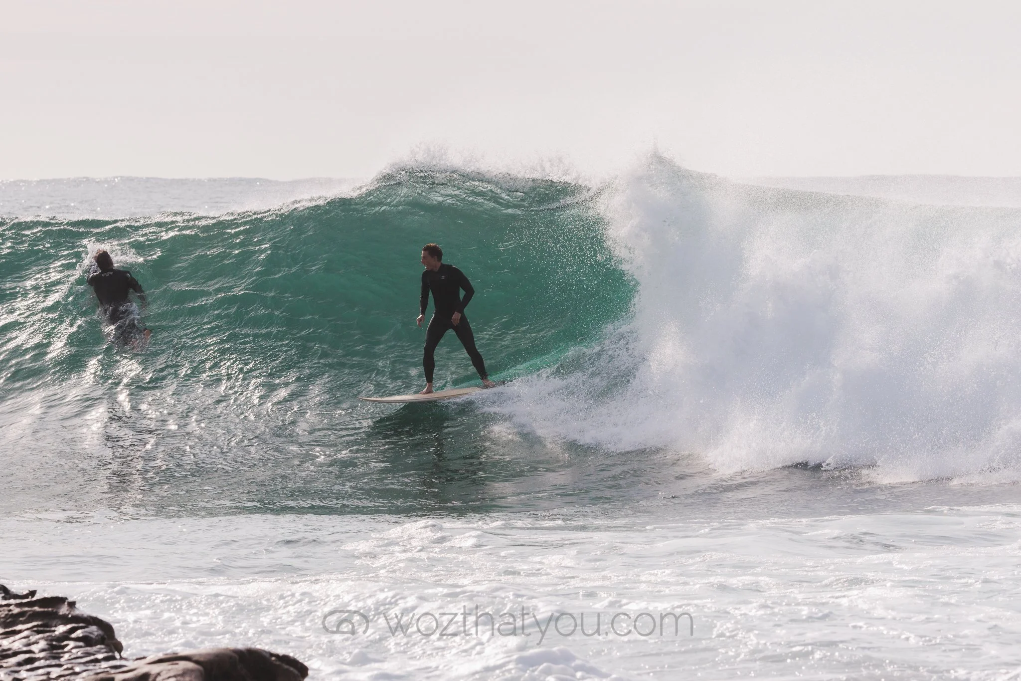 Two surfers in black wetsuits riding and approaching a large ocean wave near a rocky shoreline