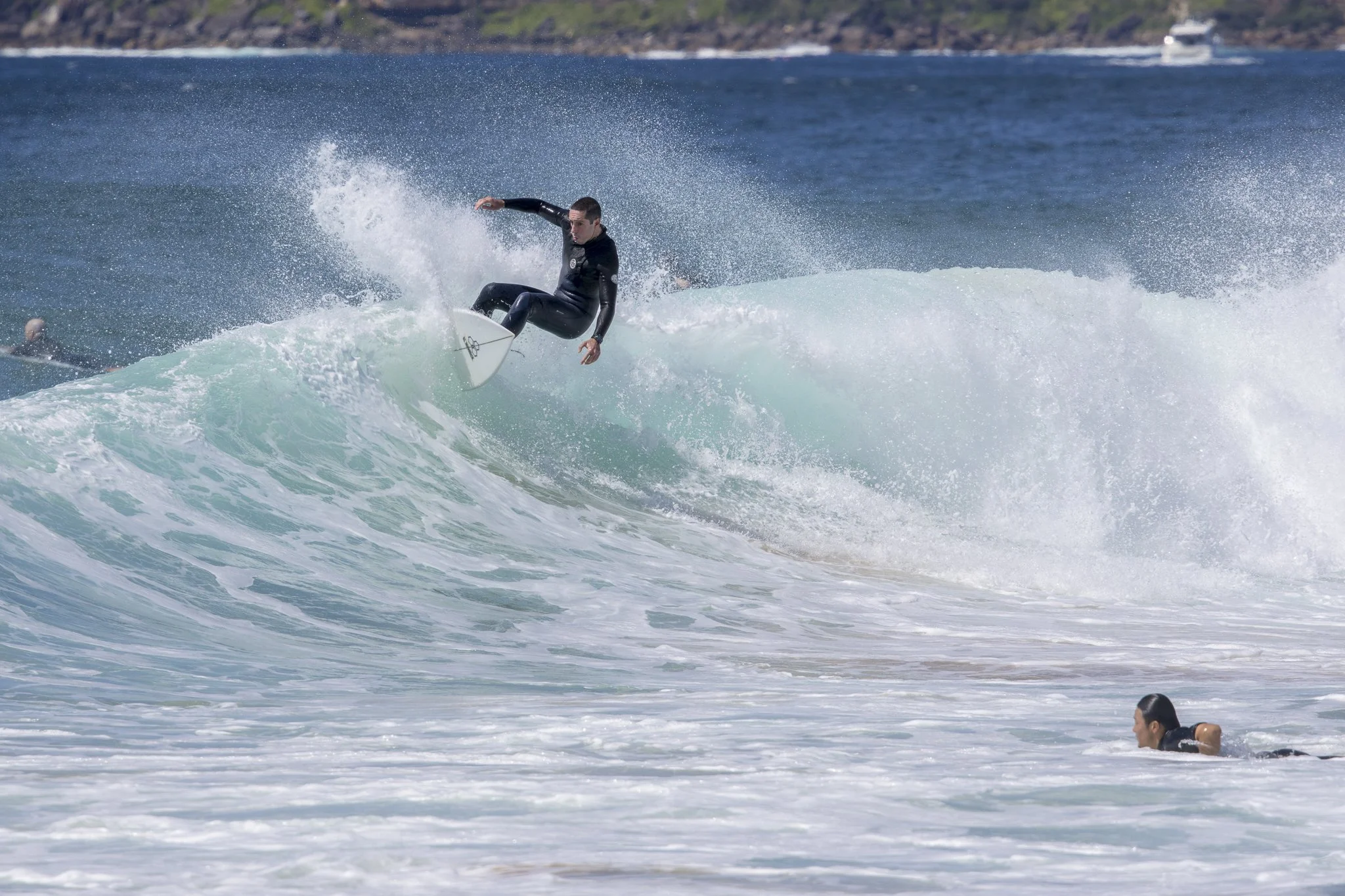 A male surfer in black wetsuit riding a cresting wave while another person swims nearby in the ocean.