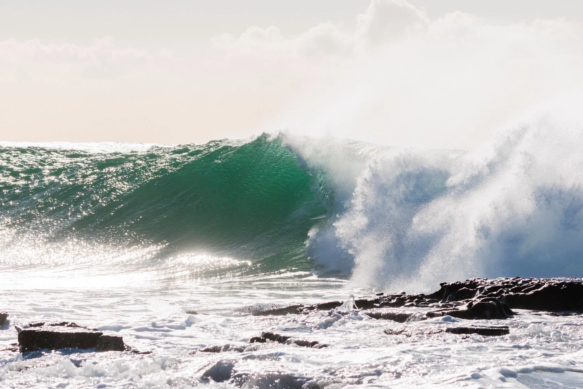 Ocean wave crashing onto rocky shore, with foam and spray in bright sunlight.