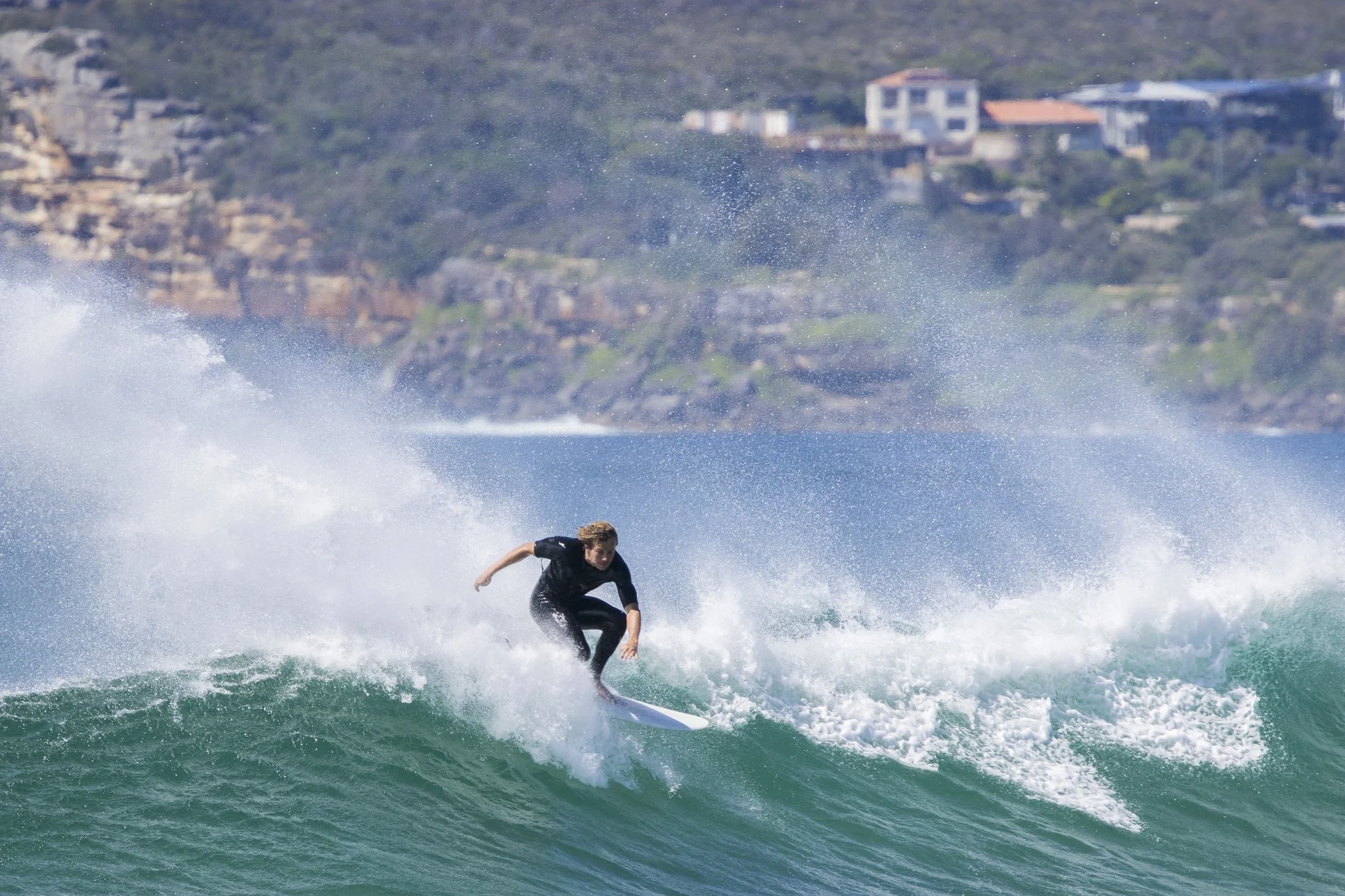 A person surfing on a wave in the ocean with a rocky coastline and houses in the background.