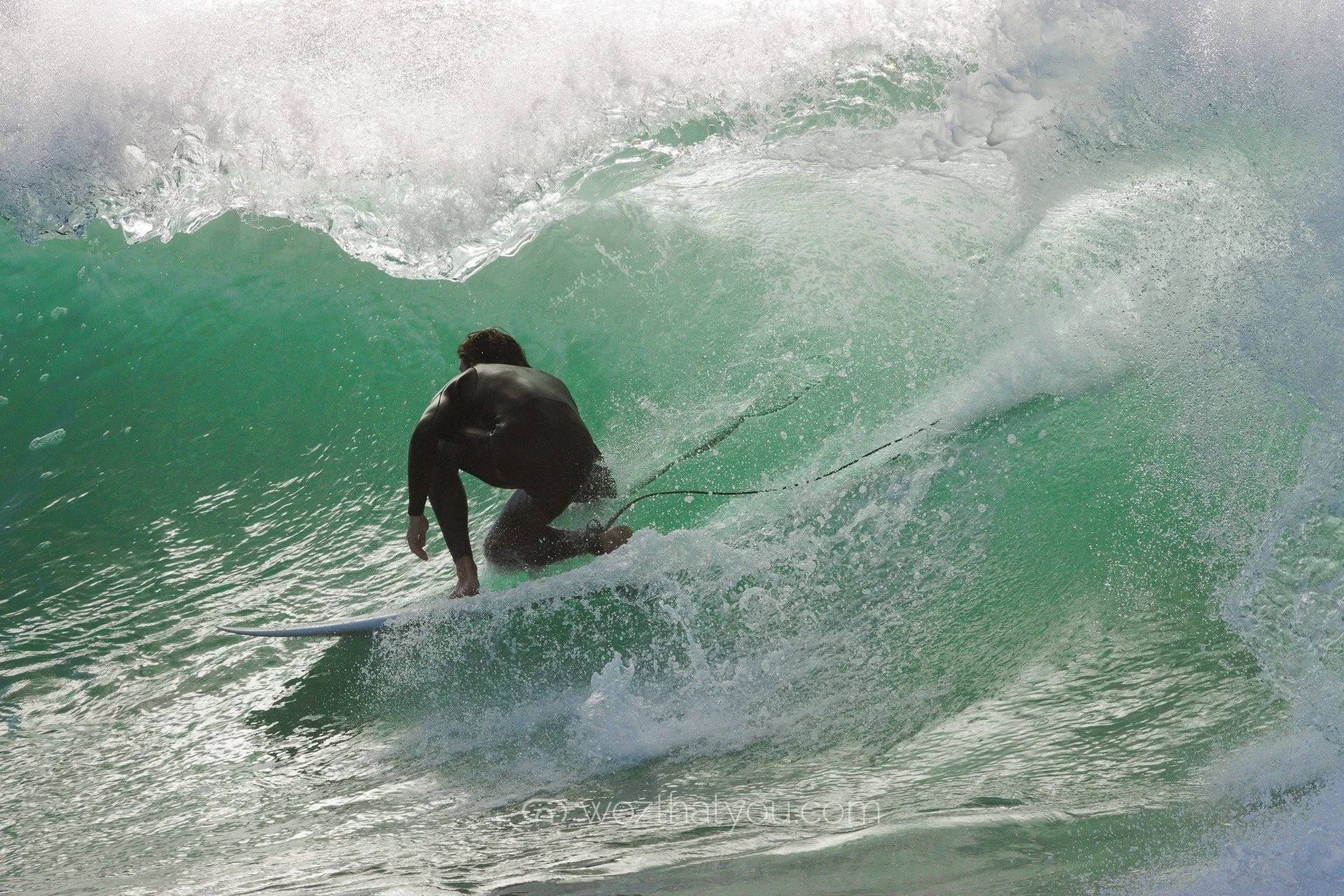 A person wearing a wetsuit is riding a surfboard inside a green wave in the ocean.