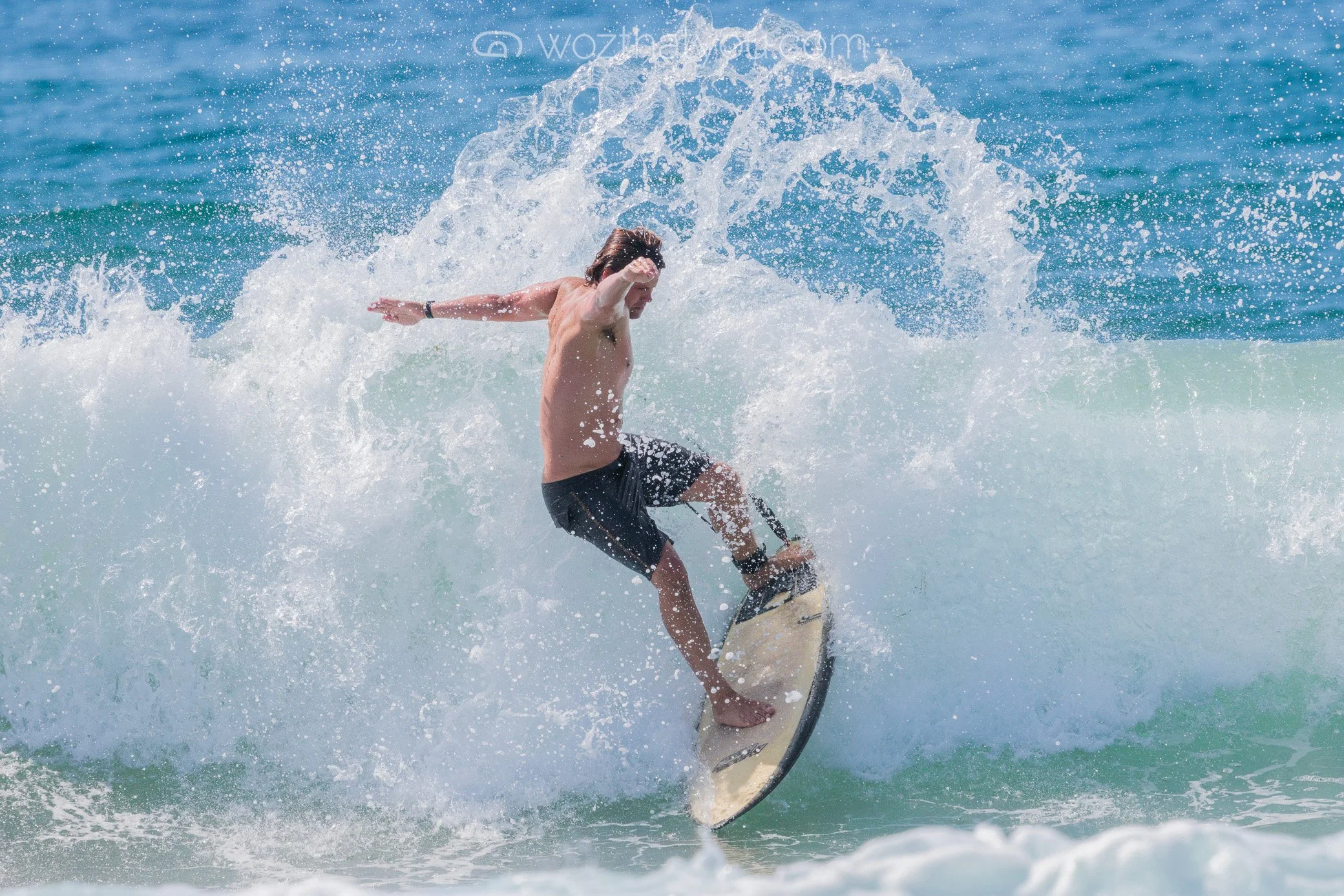 A man surfing on a wave in the ocean, wearing black shorts, with water splashing around him.