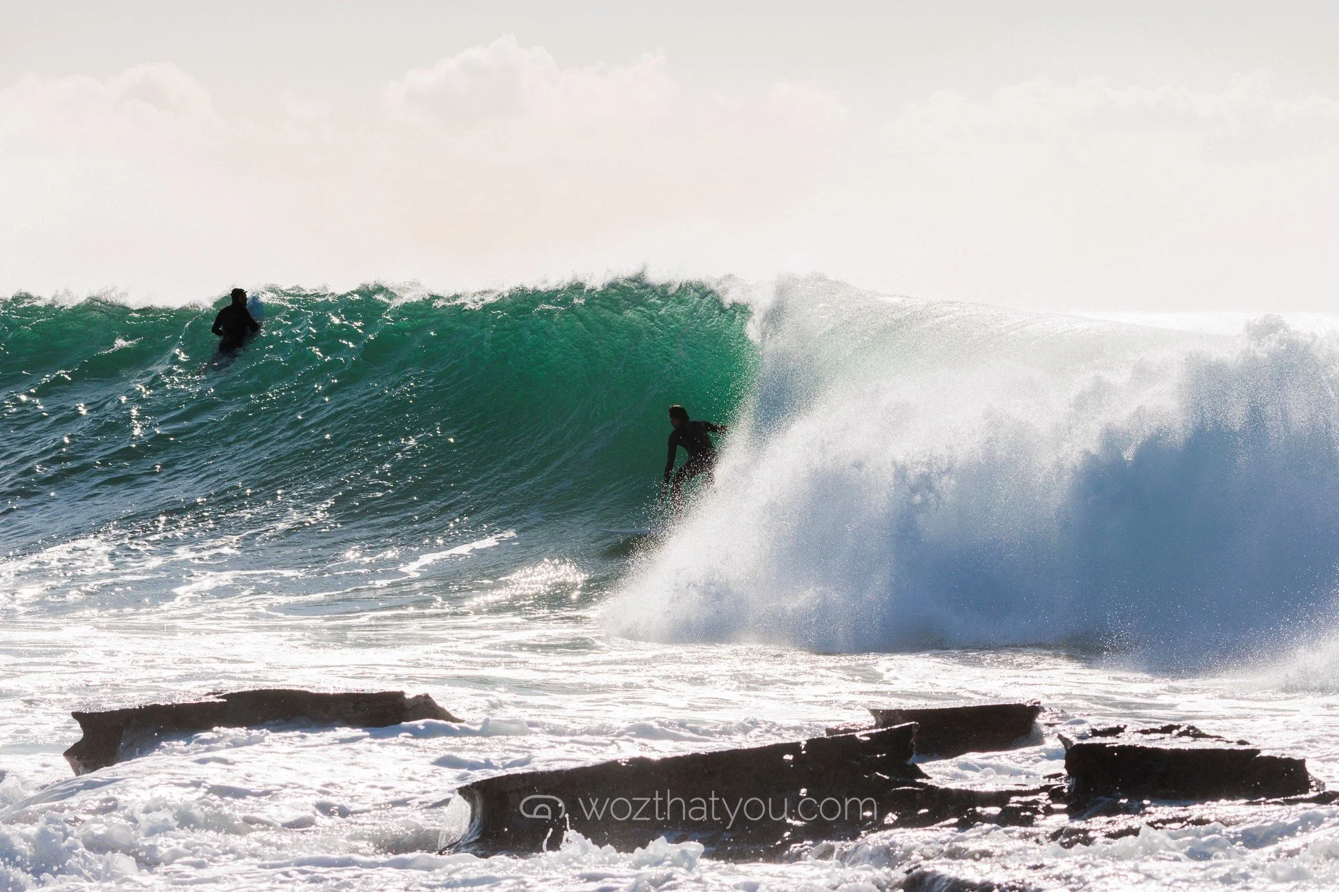 Two surfers in black wetsuits riding and waiting on a large wave near the rocky shoreline under a cloudy sky.