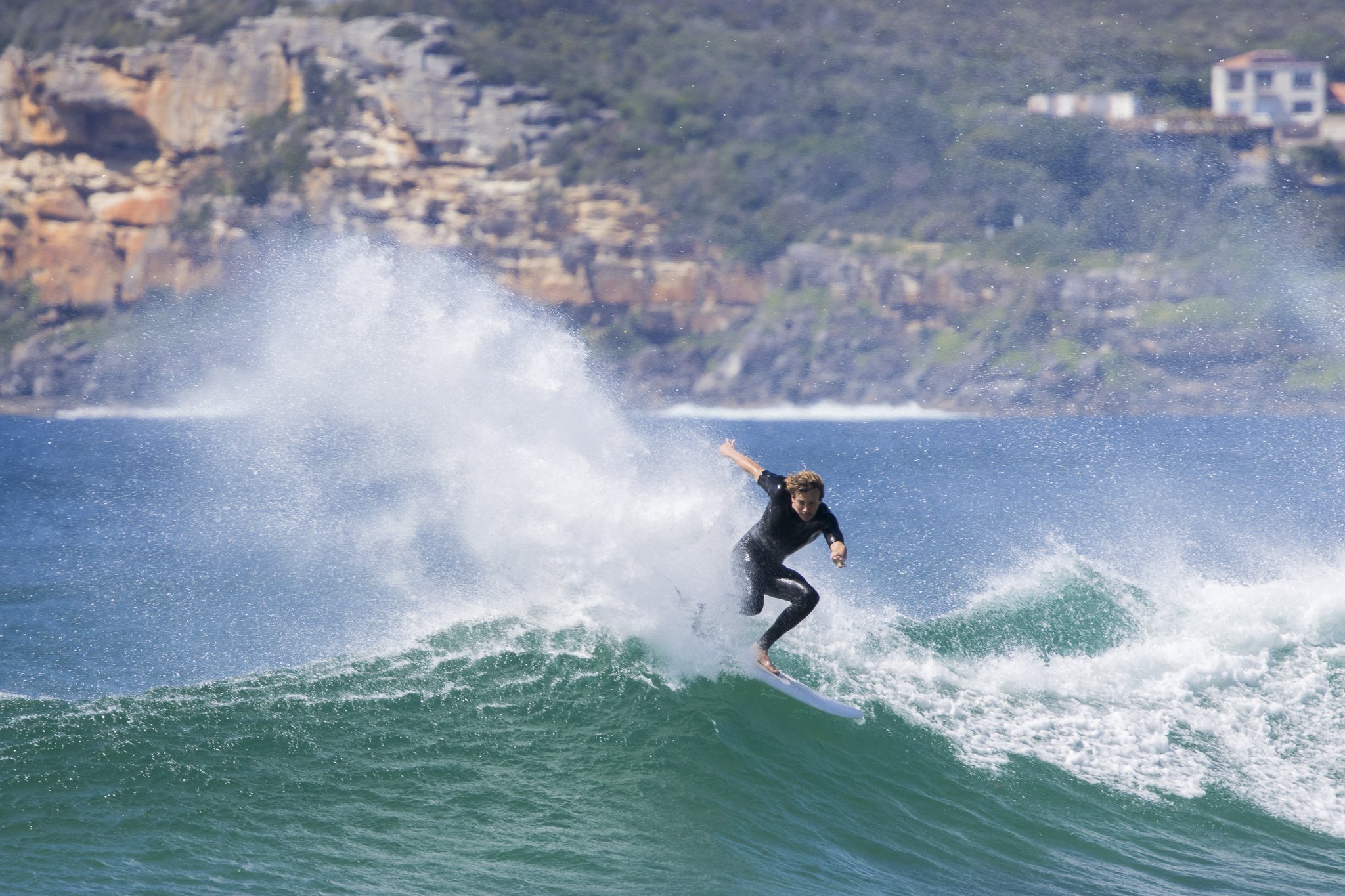 A person surfing on a wave in the ocean with a coastal hillside in the background.