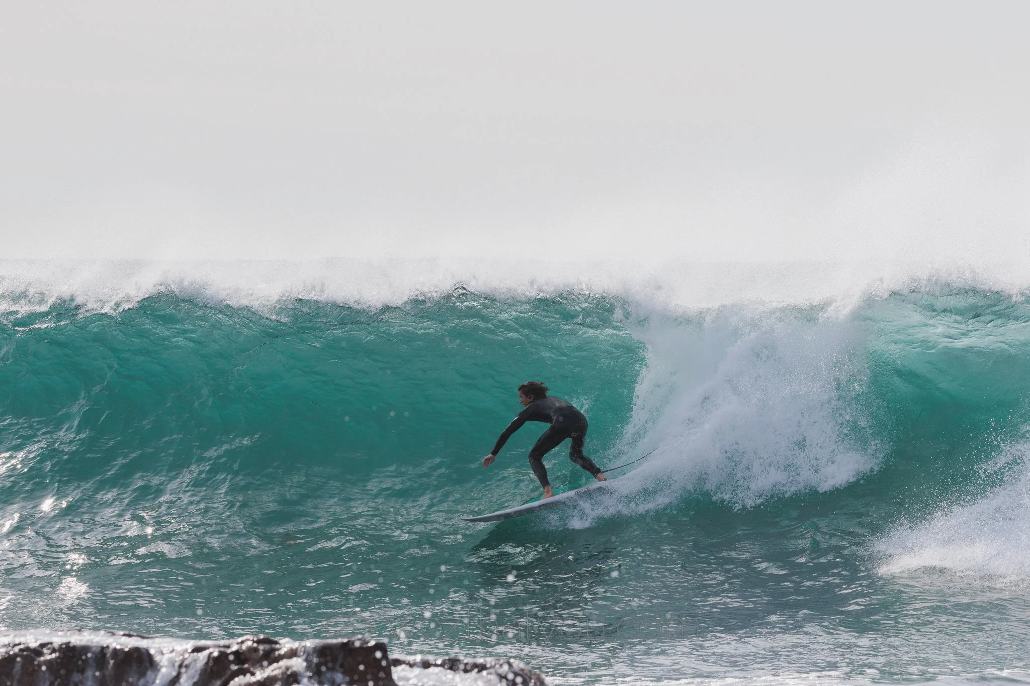 A person surfing on a large wave in the ocean, wearing a black wetsuit, with water splashing around.