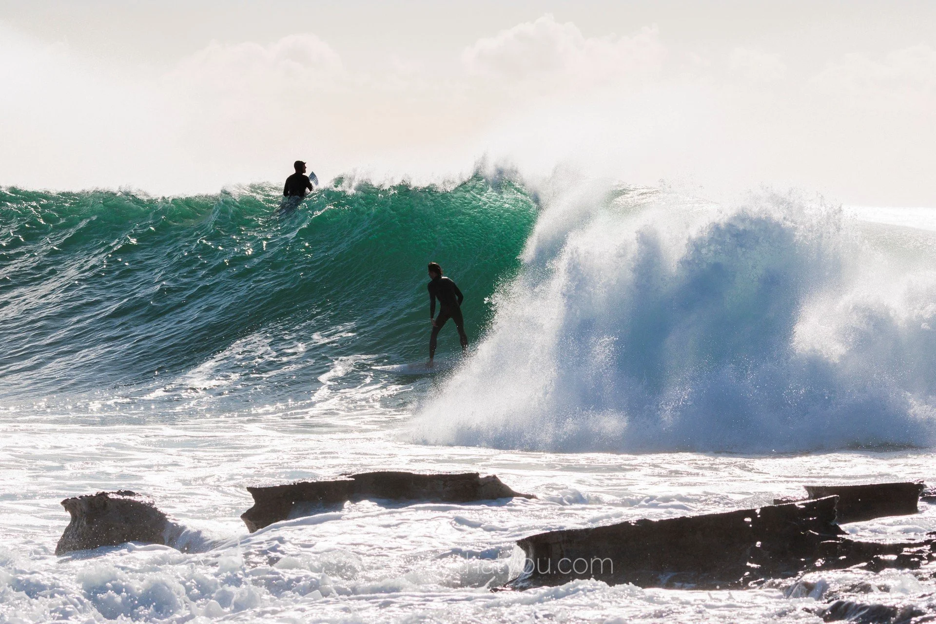 Two surfers riding a large ocean wave near rocky shoreline with spray flying off the crest.