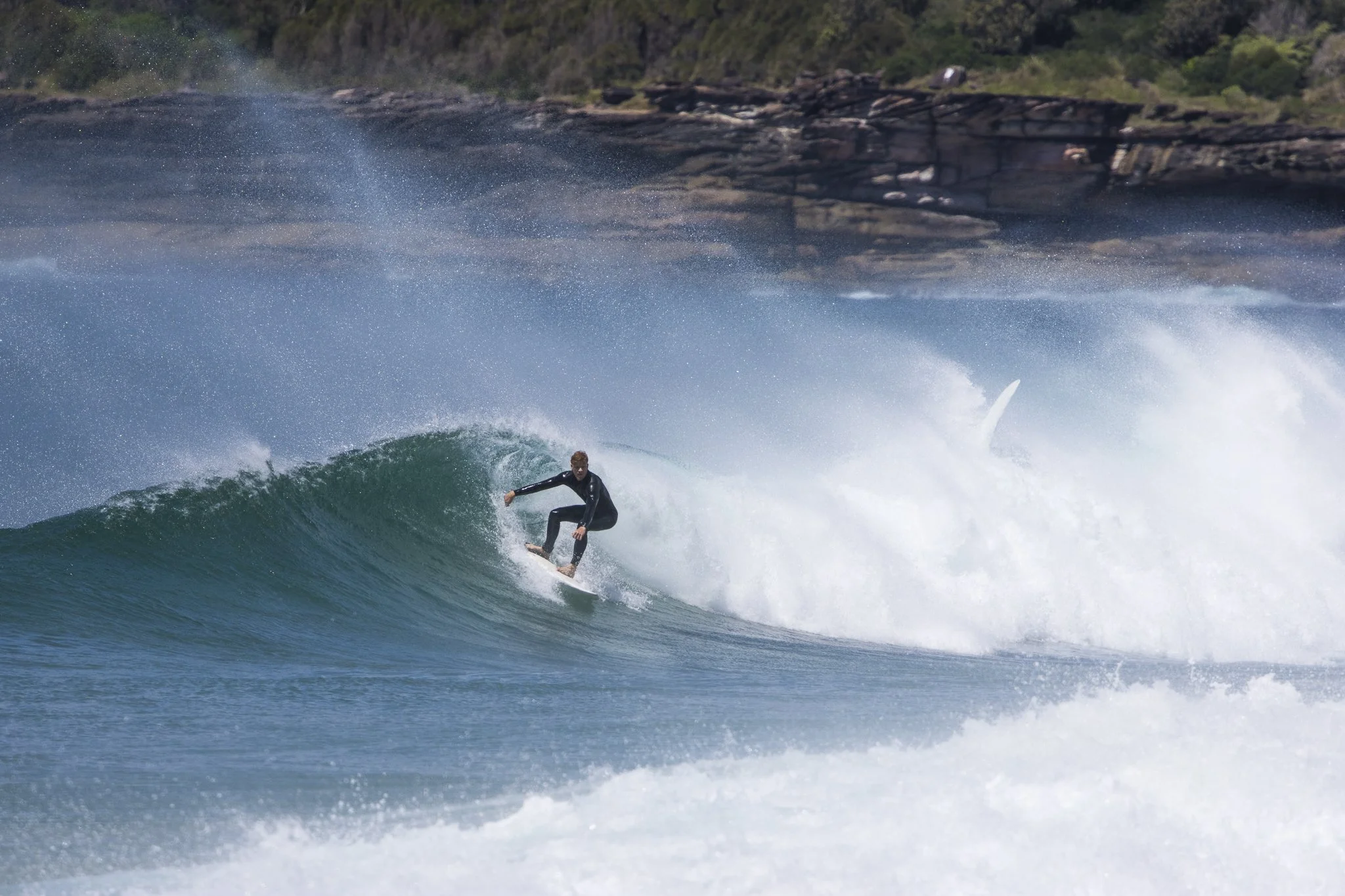 Surfer riding a wave in the ocean with rocky coastline and greenery in the background.