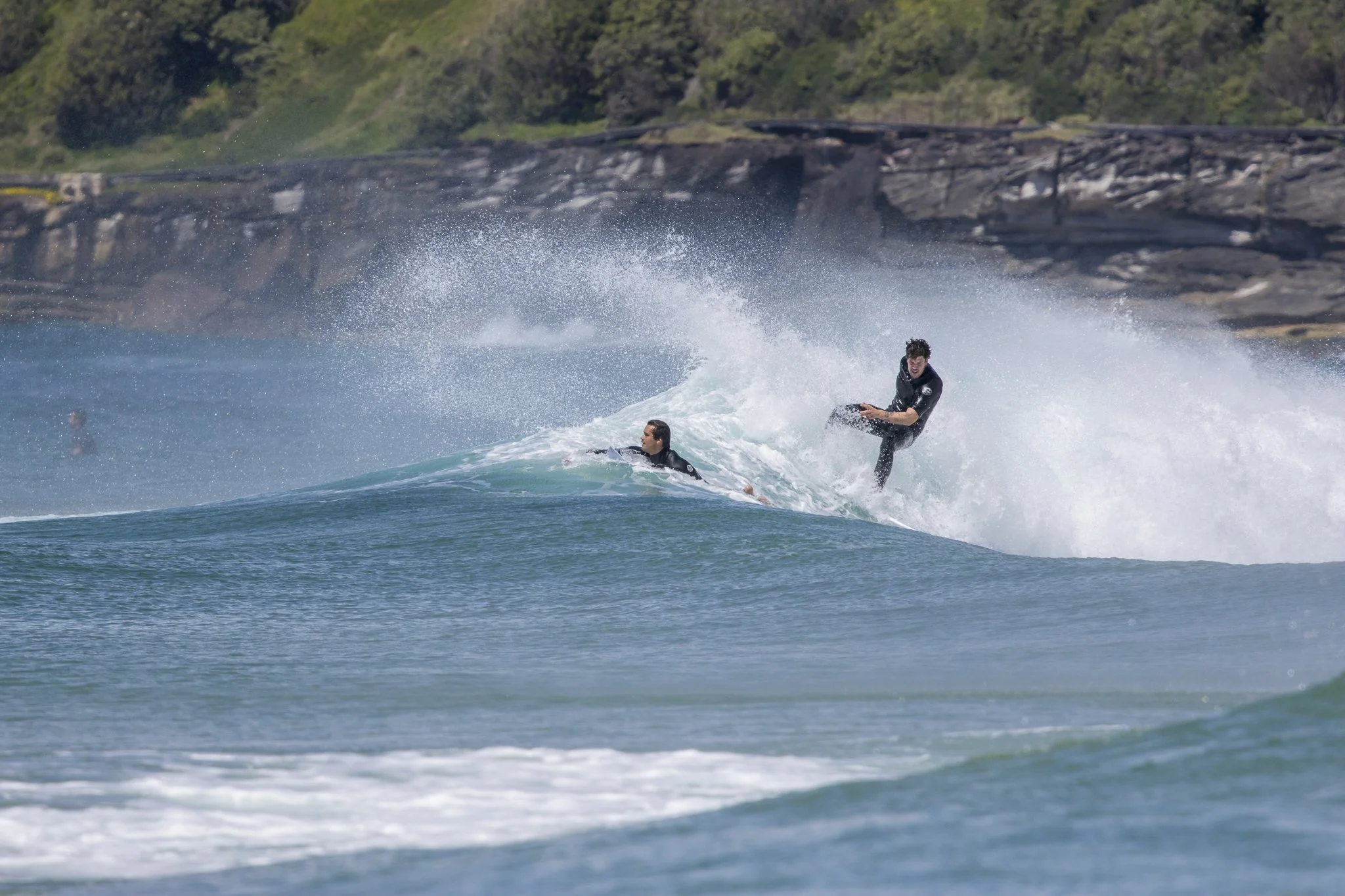 Two surfers in wetsuits riding a wave with a rocky shoreline and green vegetation in the background.