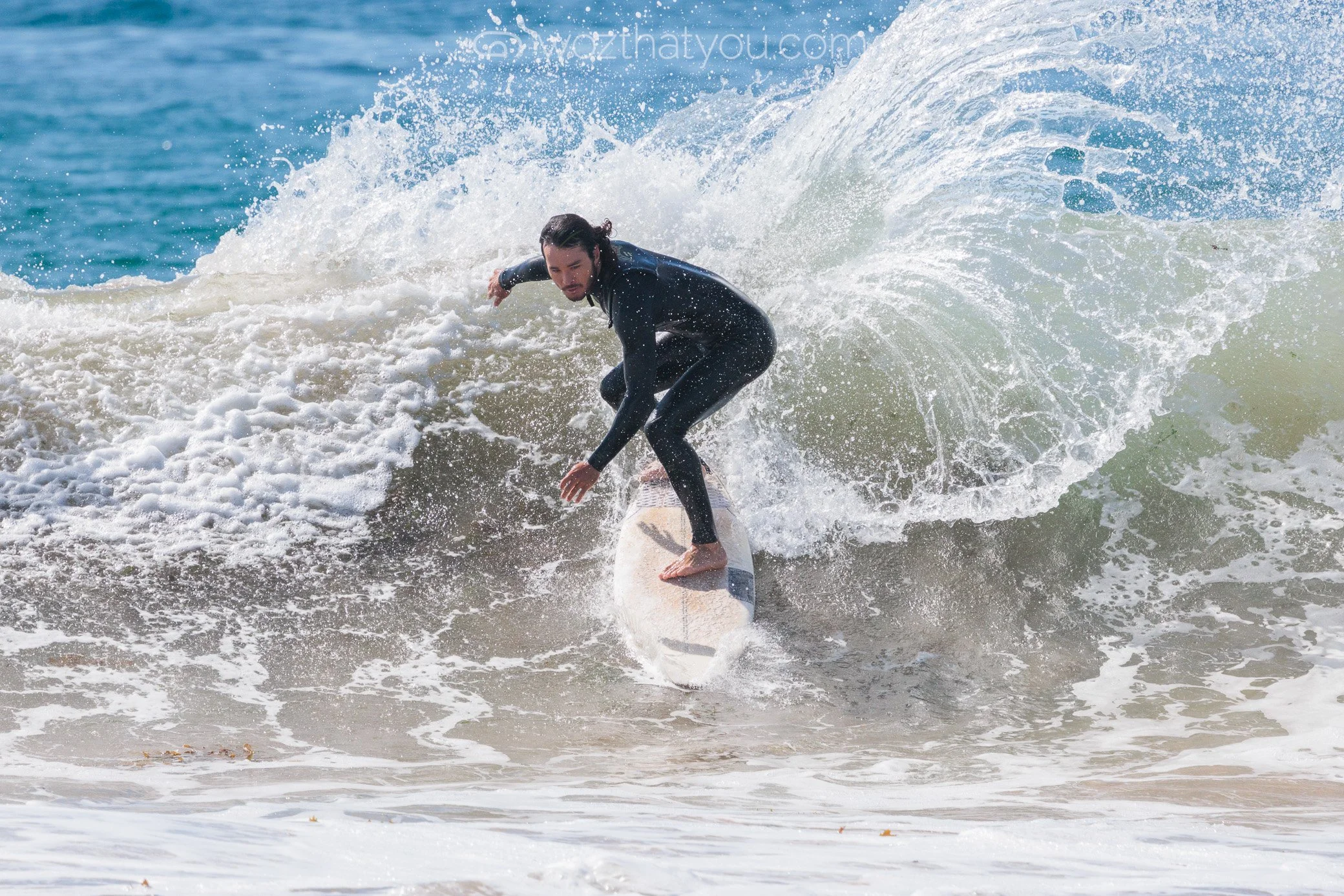 A man in a black wetsuit surfing on a white surfboard on a wave with white foam, at the beach.
