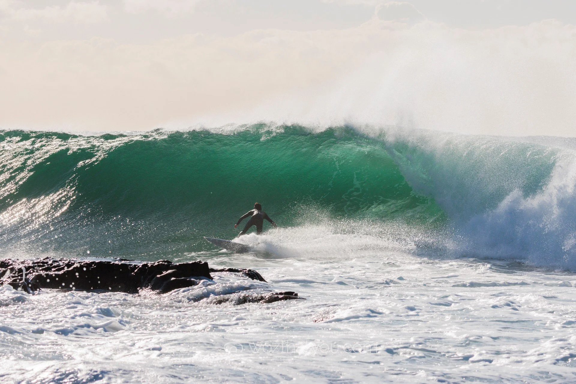 A surfer riding inside a large, green ocean wave near rocks with spray in the air during daylight.