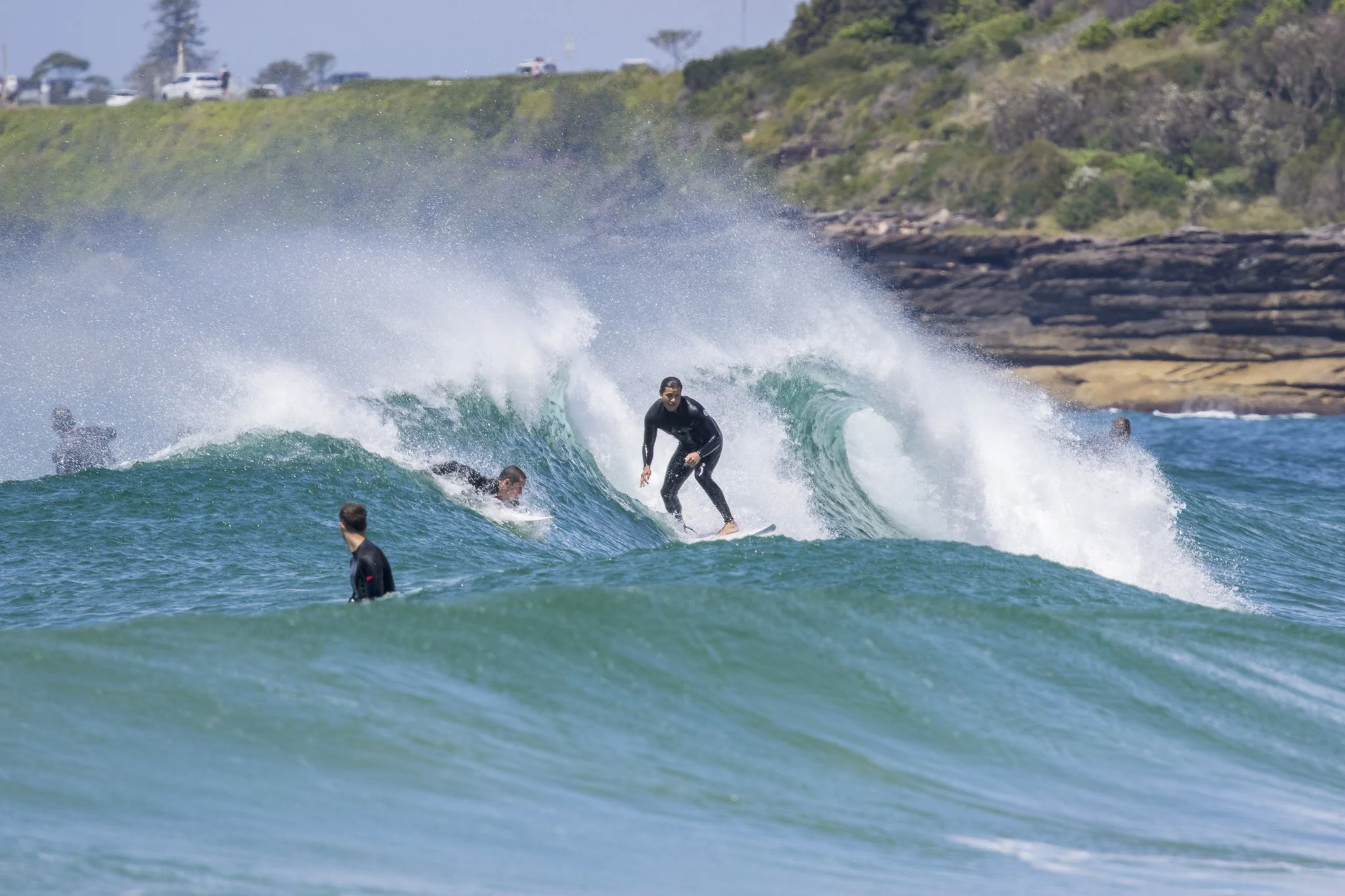 A group of surfers in wetsuits riding waves at the beach, with one surfer standing on a surfboard inside a wave and others paddling or waiting in the water.