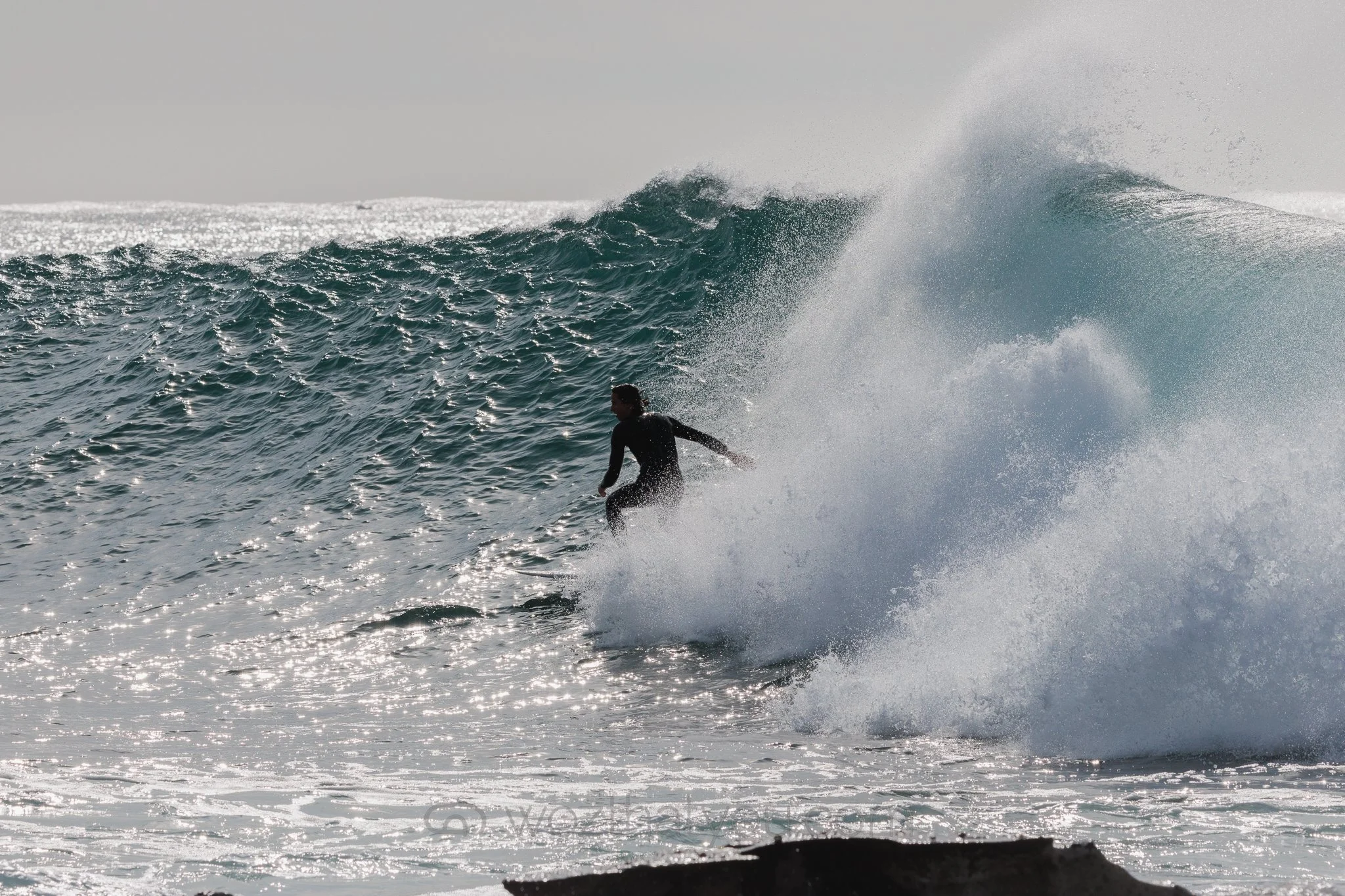 A person in a wetsuit surfing on a large ocean wave with sunlight reflecting off the water.