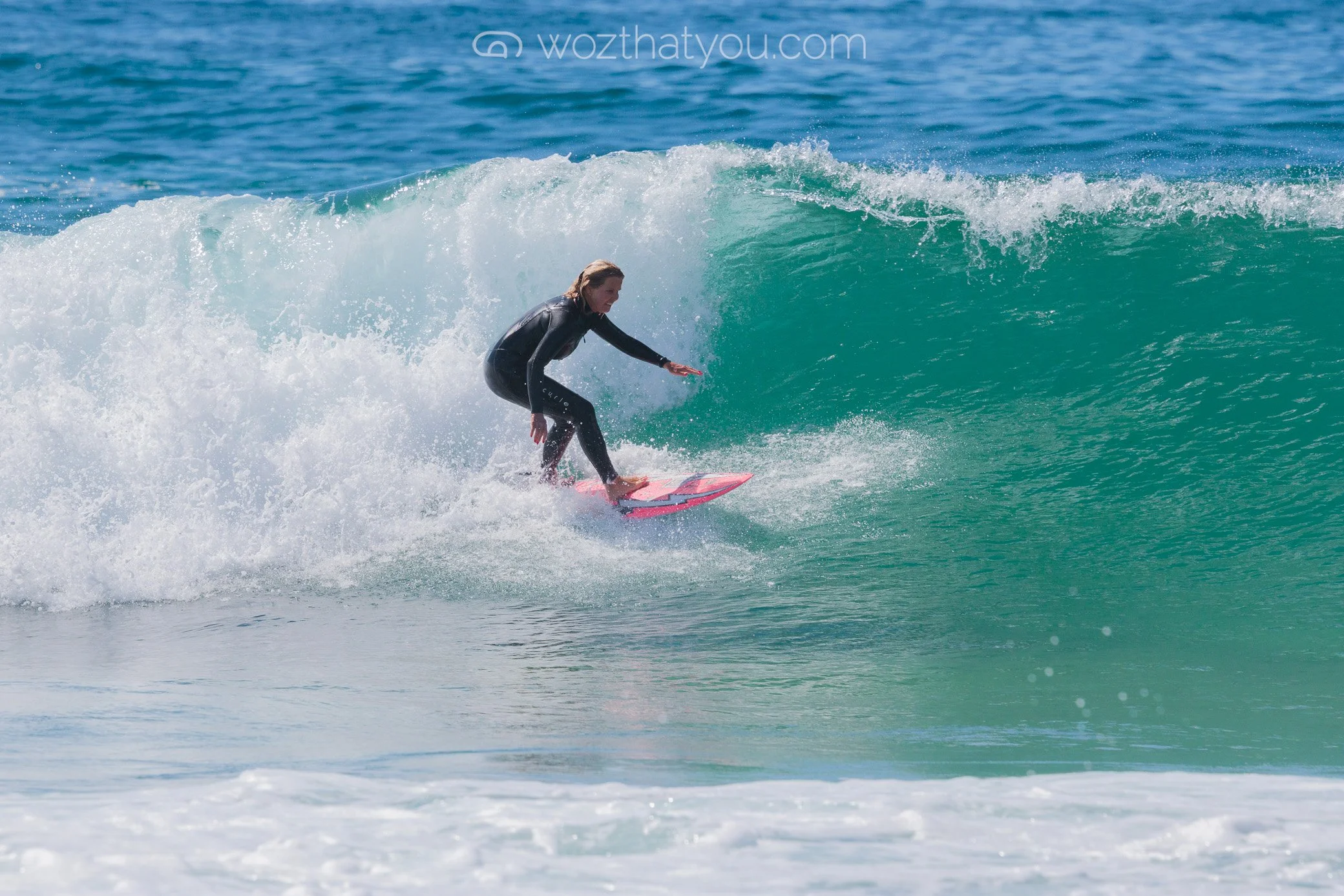 A woman surfing on a pink surfboard in the ocean, riding inside the wave with water spray.