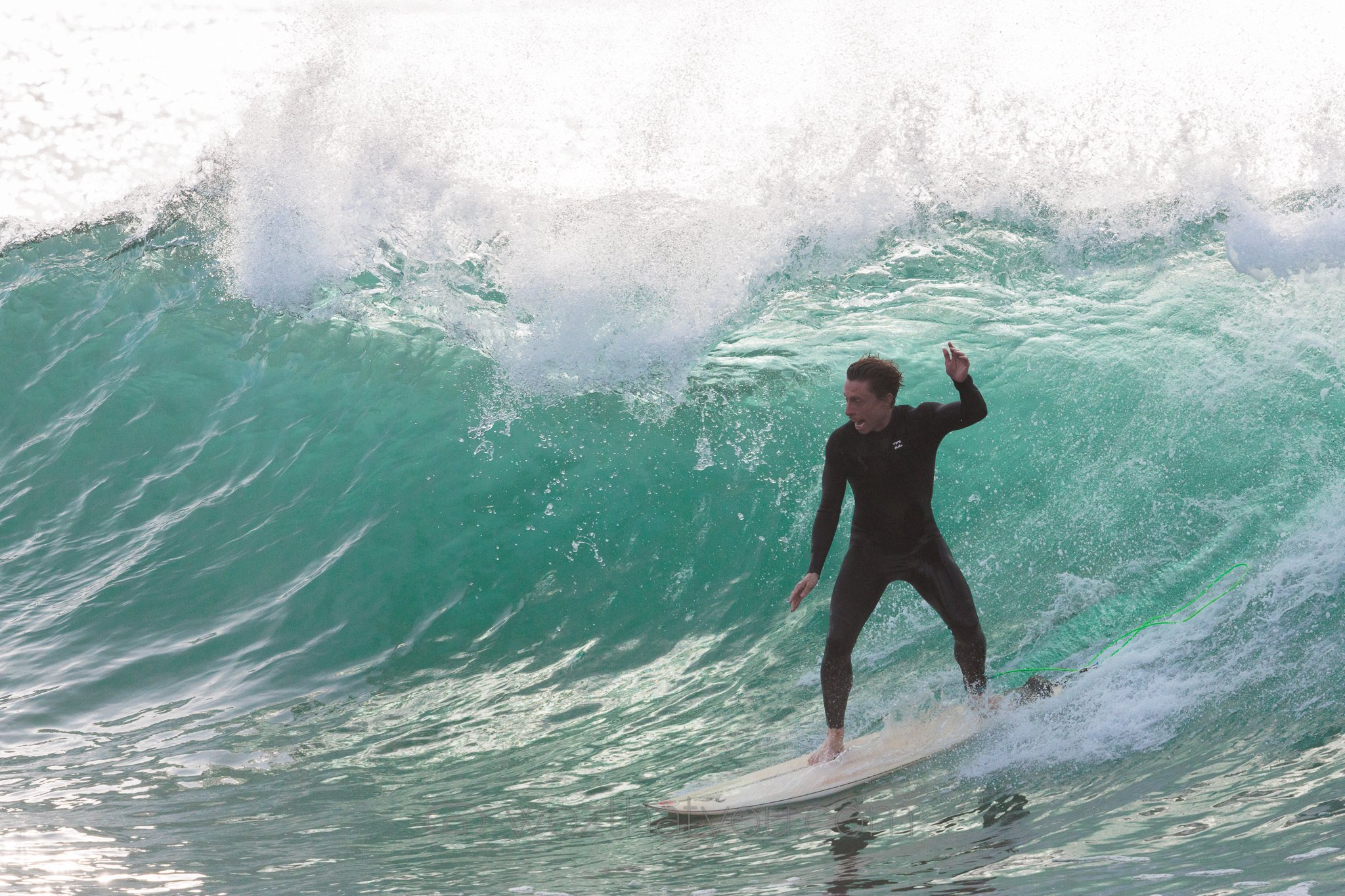 A person in a black wetsuit surfing on a white surfboard on a large green wave with white water spray.