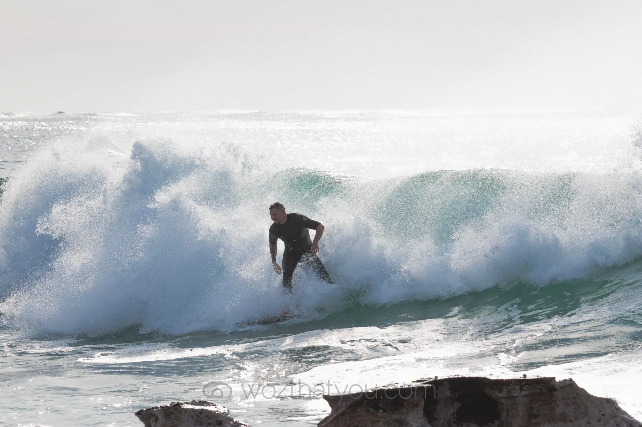 Person surfing on a wave at the beach with rocks visible in the foreground.