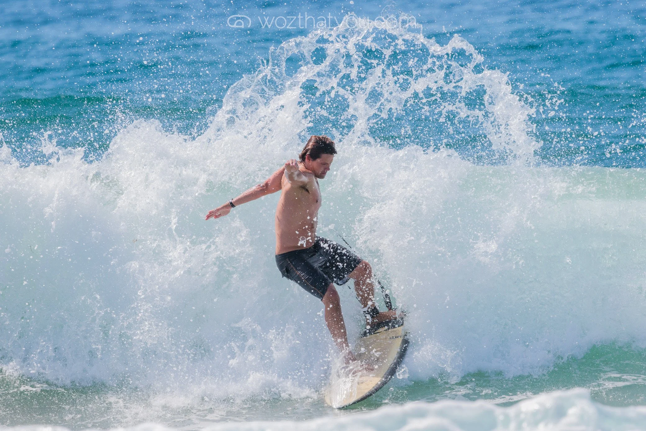 A man surfing on a wave in the ocean, shirtless and wearing black shorts.