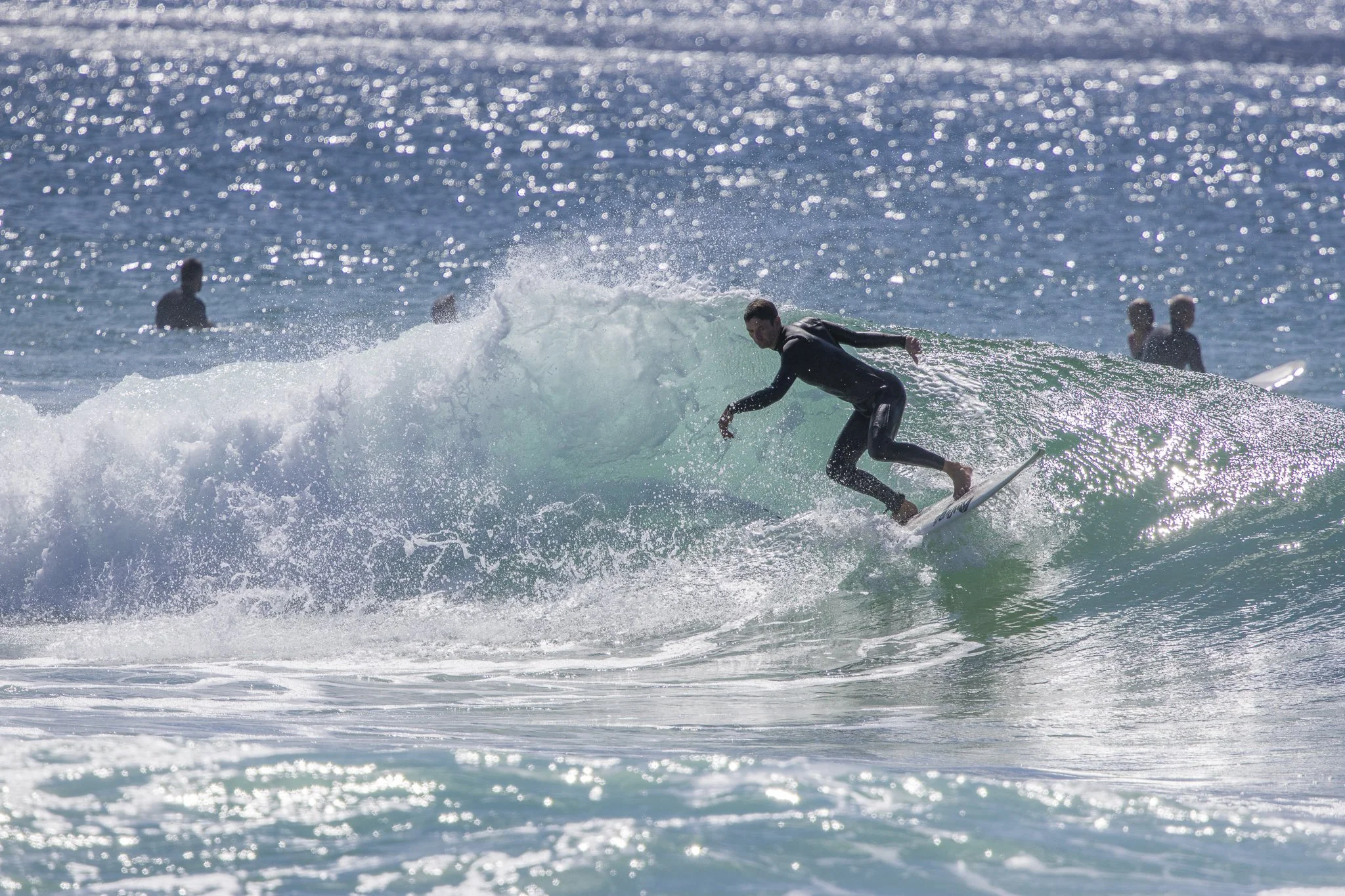 A surfer in a black wetsuit riding a wave in the ocean with sparkling water in the background and other surfers floating in the distance.